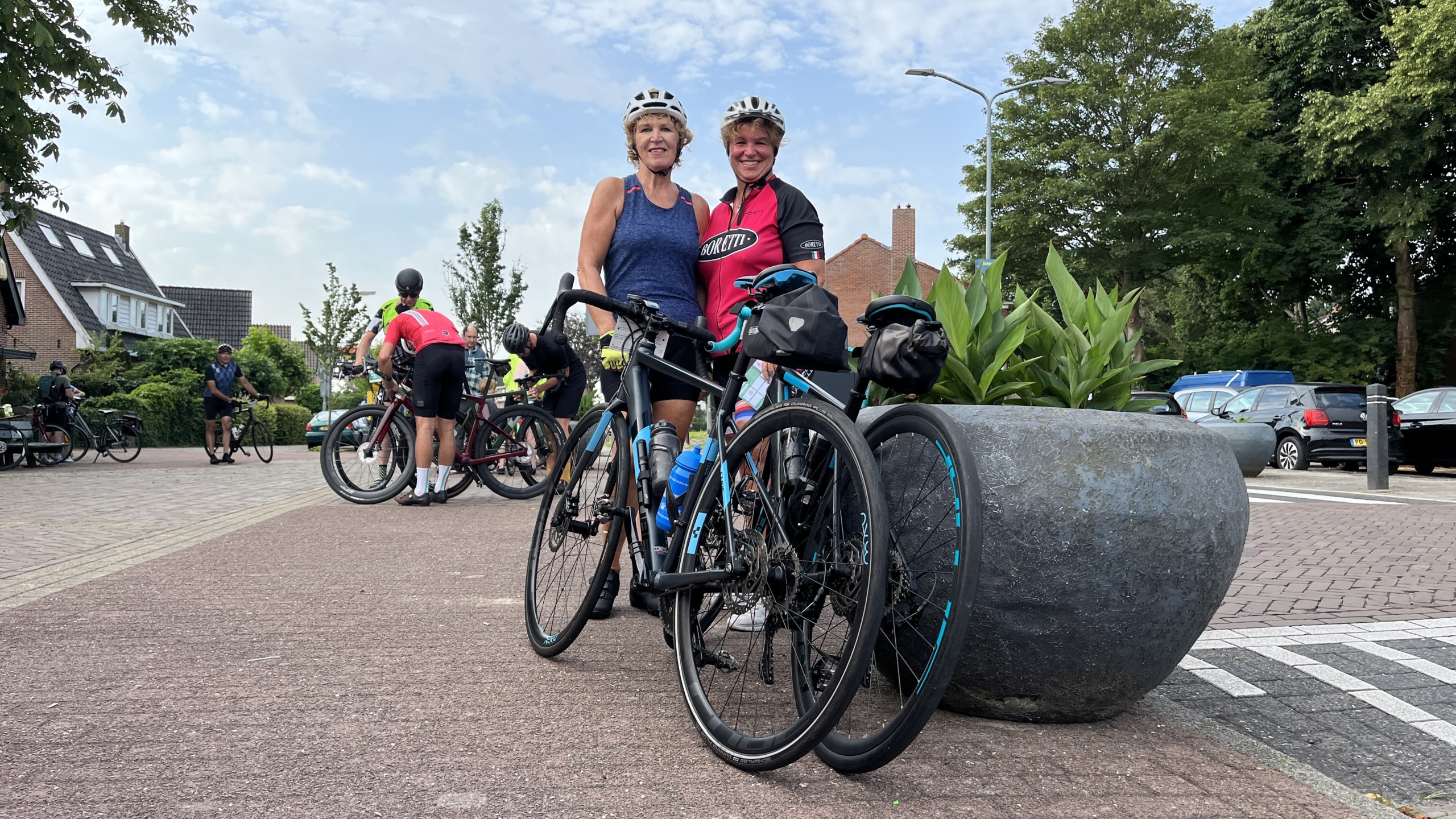 Twee vrouwen in wielerkleding poseren met hun fietsen op een zonnige dag, omringd door andere fietsers op een plein met plantenbak en geparkeerde auto's op de achtergrond.