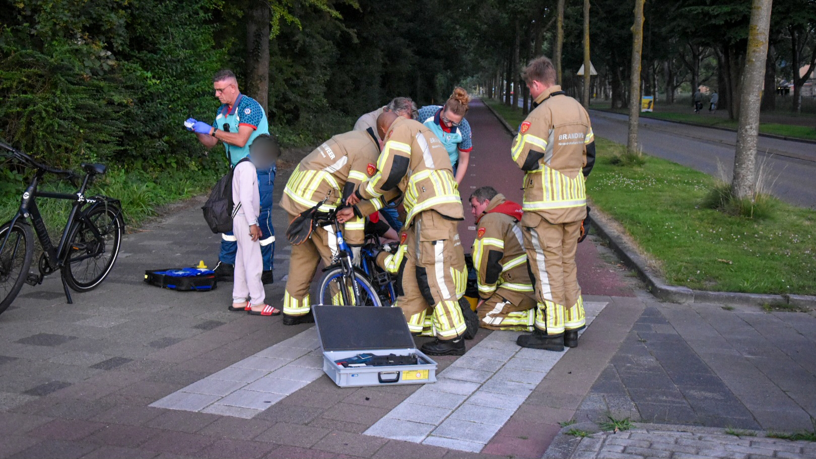 Hulpverleners, waaronder brandweerlieden in uniform, verlenen hulp bij een fietsongeluk op een trottoir. Een ambulancebroeder staat met een kind en medische apparatuur is aanwezig op de grond. Omgeving met bomen en een fietspad zichtbaar.