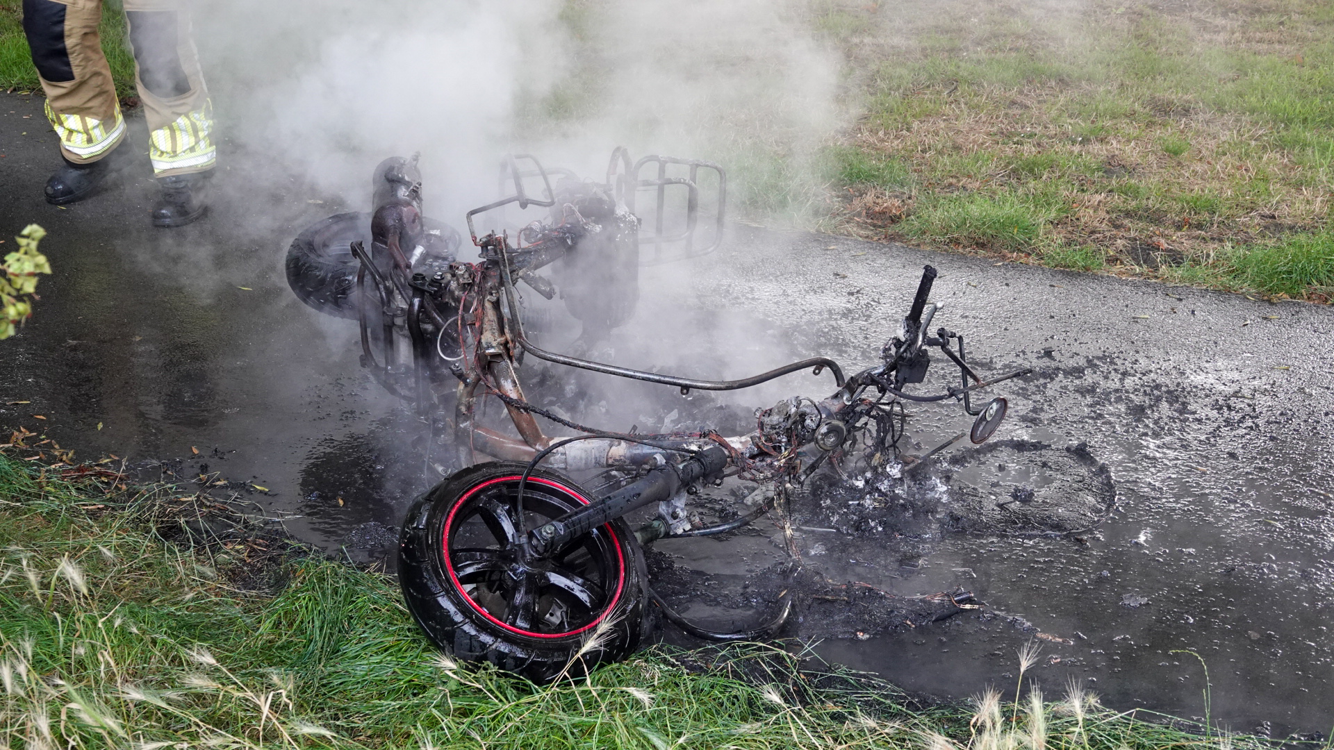 Een verbrande fiets, grotendeels gesmolten en zwartgekoold, ligt op een nat trottoir met rook die er vanaf komt, terwijl een persoon in een brandweeruniform naast de fiets staat.