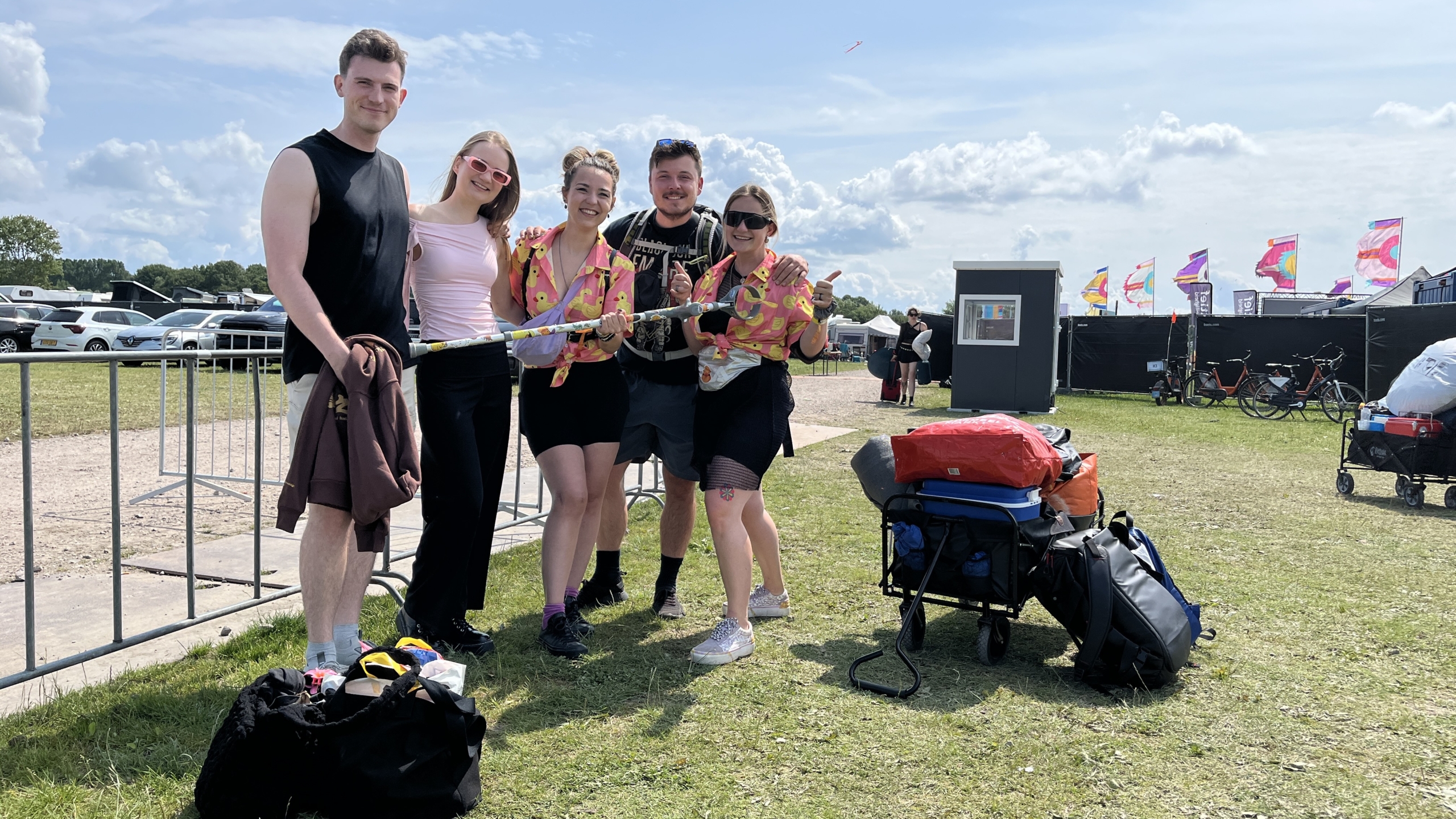 Vijf mensen poseren glimlachend voor de camera op een grasveld bij een festival, omringd door kampeeruitrusting en trolly's; op de achtergrond zijn auto's, een gebouw en kleurrijke vlaggen zichtbaar onder een deels bewolkte lucht.
