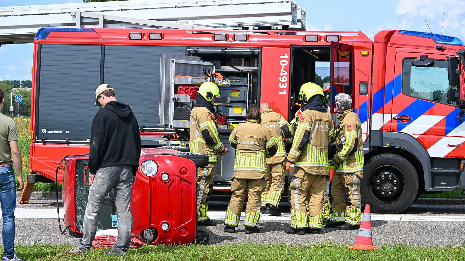 Brandweerlieden in uniform staan bij een omgevallen rode auto naast een brandweerwagen, terwijl enkele omstanders toekijken langs de weg.