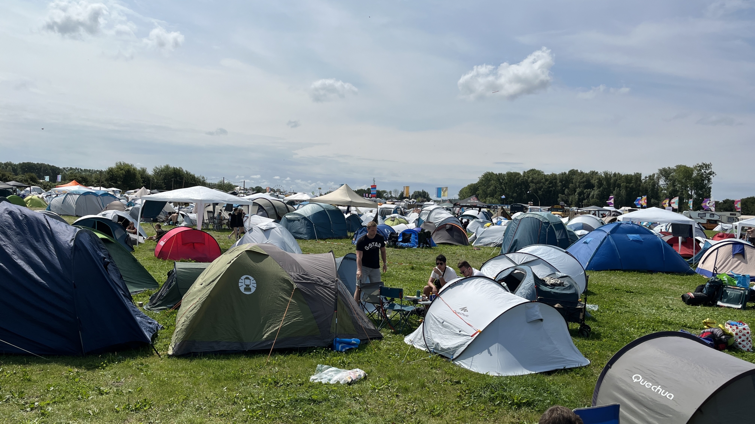 Veelzijdig kampeerterrein bij een festival, vol met kleurrijke tenten en enkele mensen die samen genieten, omgeven door een open veld en een heldere lucht.