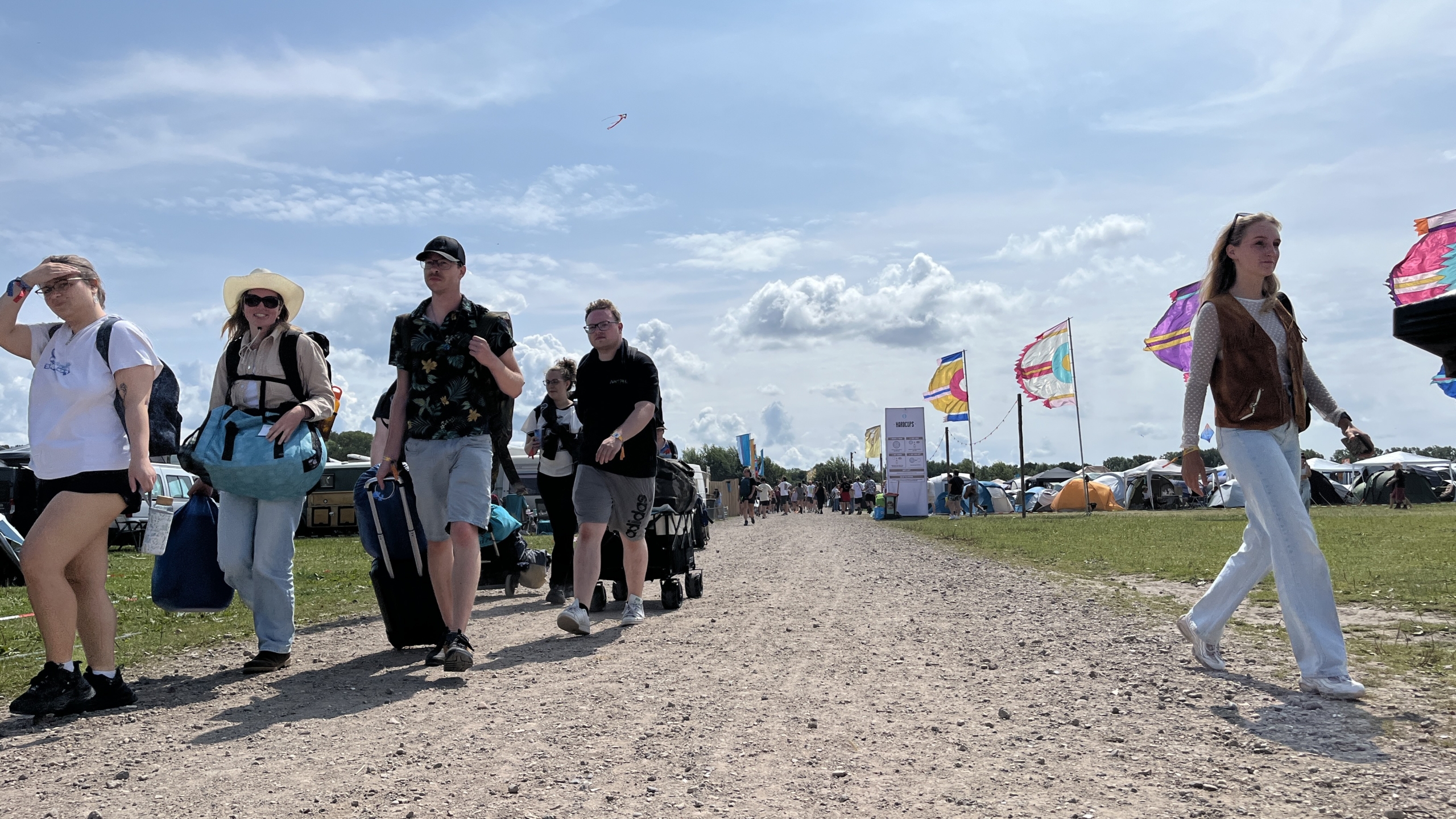 Groep mensen loopt over een stoffig pad op een festivalterrein, met tenten en gekleurde vlaggen op de achtergrond onder een zonnige hemel.