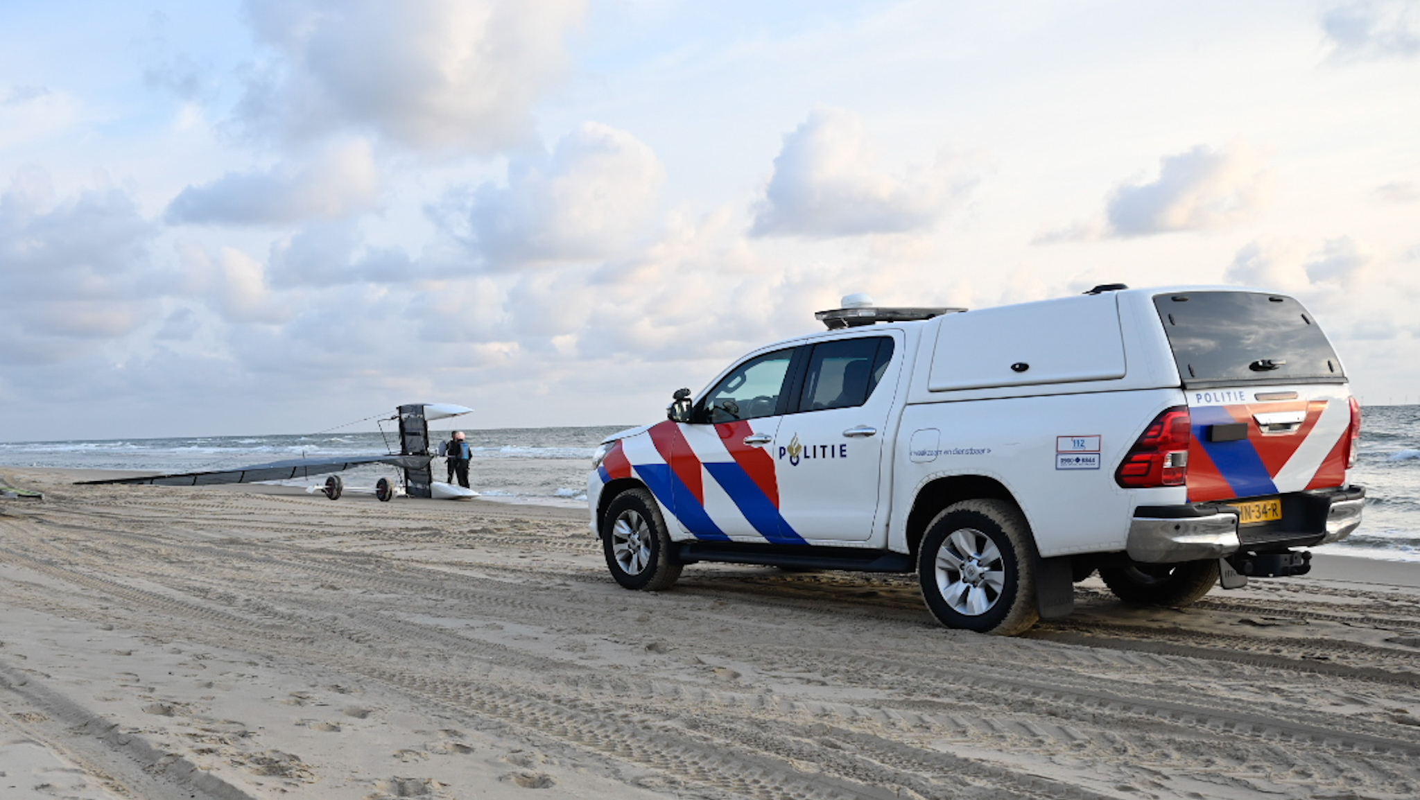 Een politieauto staat geparkeerd op een zandstrand naast een glider-vliegtuigje dat aan de kust ligt, met een persoon die ernaar kijkt. De lucht is bewolkt en de zee is op de achtergrond te zien.