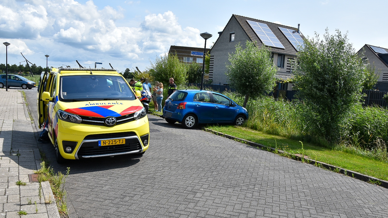 Gele ambulance en een blauw voertuig langs de weg bij een woning met zonnepanelen; mensen en hulpverleners zijn aanwezig.
