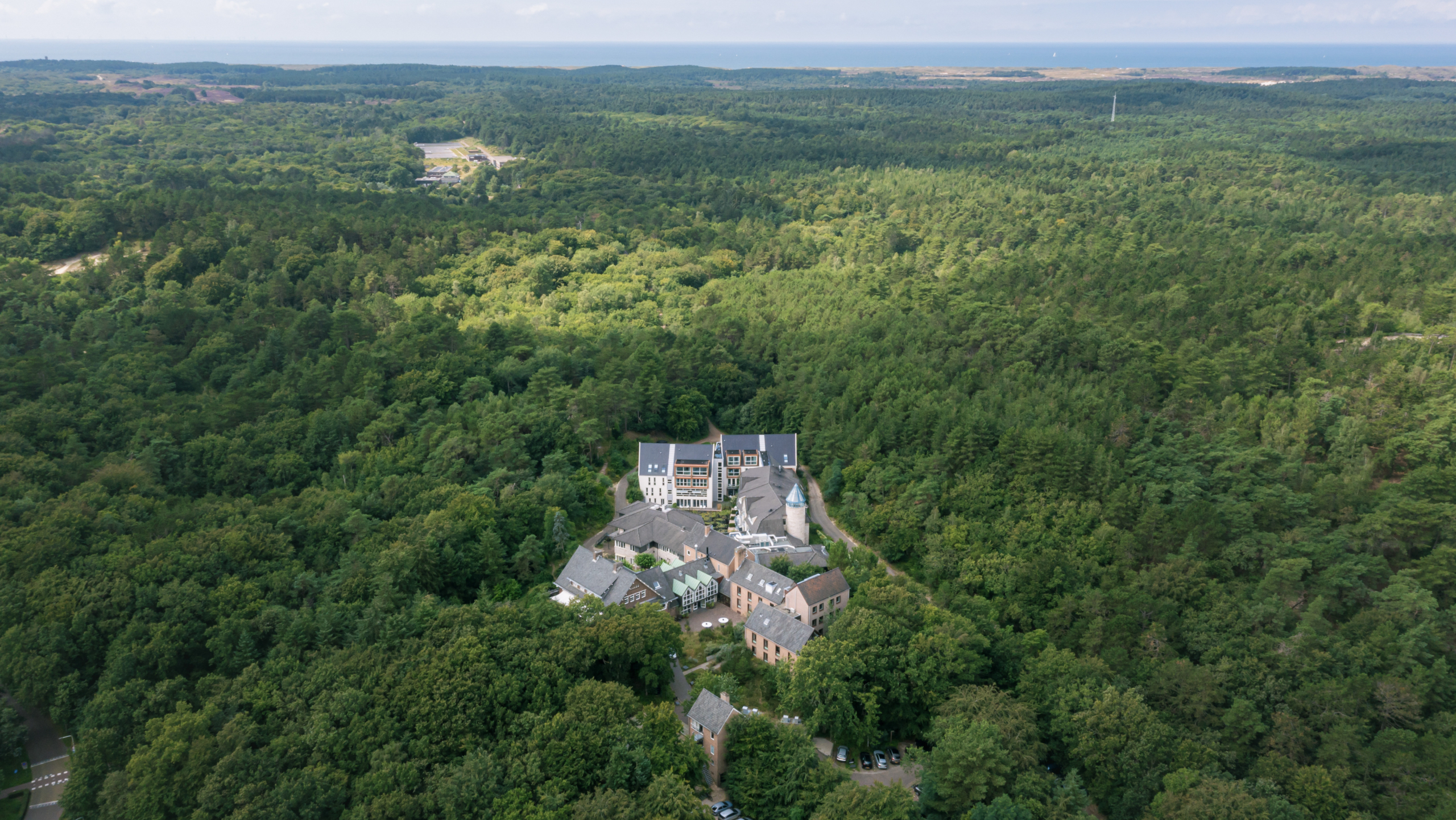 Luchtfoto van een kasteelachtig gebouw omgeven door dichte groene bossen, met een uitgestrekt landschap op de achtergrond dat zich uitstrekt tot aan de horizon.
