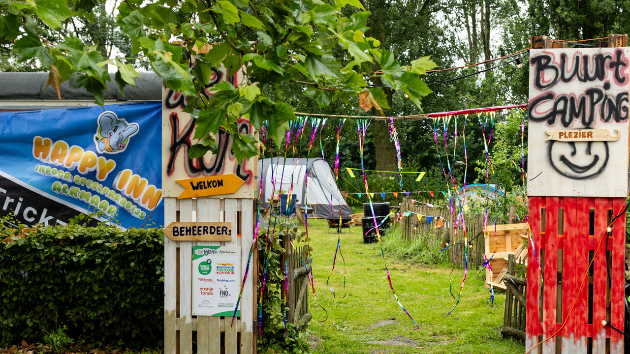 Ingang van een buurtcamping met borden die "Welkom" en "Beheerder" aangeven, kleurrijke linten en een tent op de achtergrond, omgeven door groene bomen en struiken.