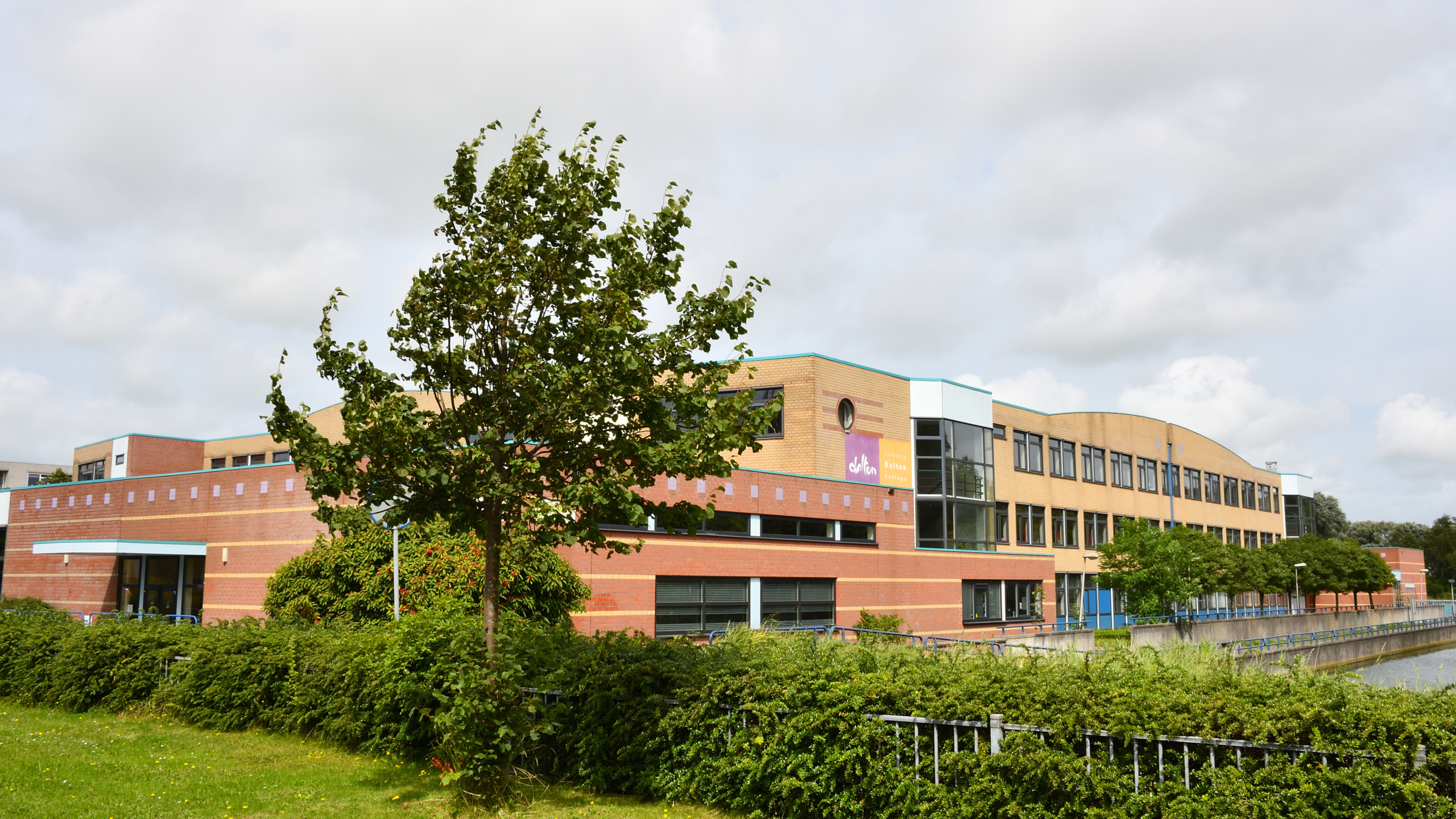 Een modern gebouw van baksteen en glas, omgeven door een groen landschap met een boom op de voorgrond en een gracht aan de zijkant, onder een bewolkte hemel.
