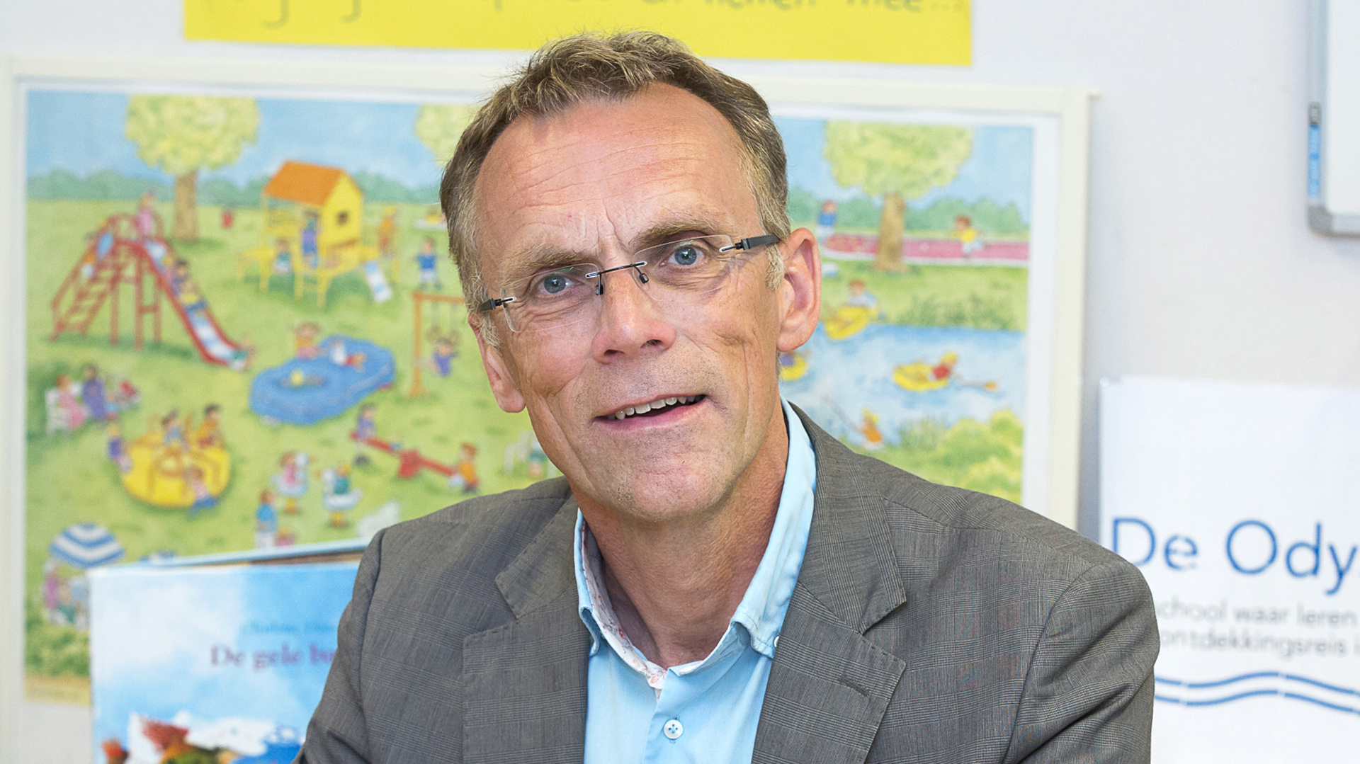 Man met bril en grijs haar draagt een grijs pak en lichtblauw overhemd, zittend voor een kleurrijk schoolbord met afbeelding van een speeltuin.