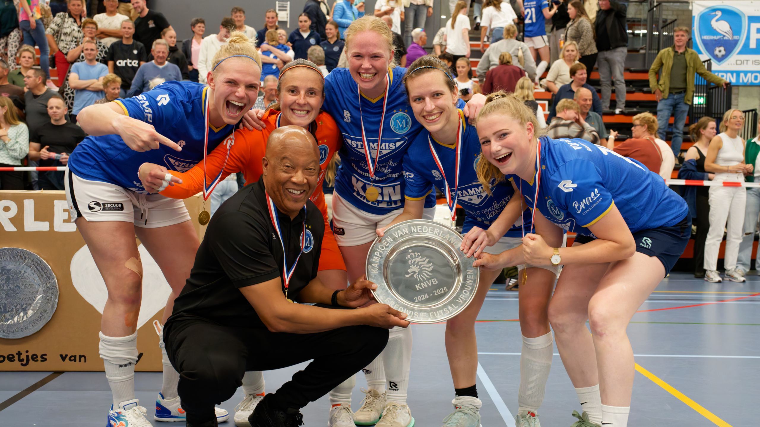 Een groep van vijf vrouwen in blauwe sportkleding en een man in zwarte kleding poseren trots met een kampioenschaal in een sporthal, omringd door juichende toeschouwers.