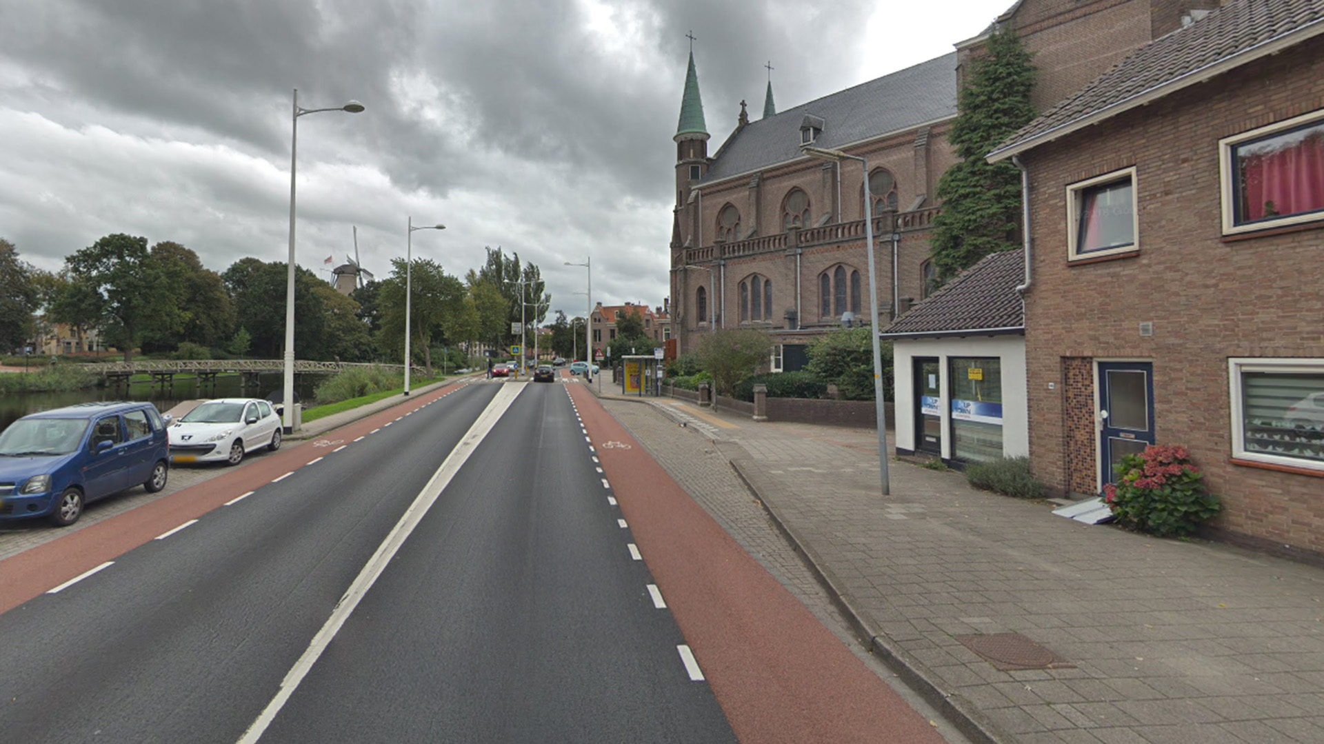Straatbeeld met een kerk met een hoge torenspits aan de rechterkant en enkele geparkeerde auto's aan de linkerkant van de weg, met een fietspad naast het verkeer. Op de achtergrond is een windmolen te zien en de lucht is bewolkt.