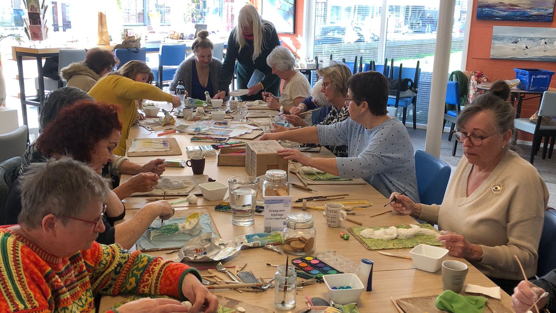Groep mensen zit aan een tafel in een creatieve workshop in een lichte ruimte, omringd door verschillende kunstmaterialen zoals verf en papier.