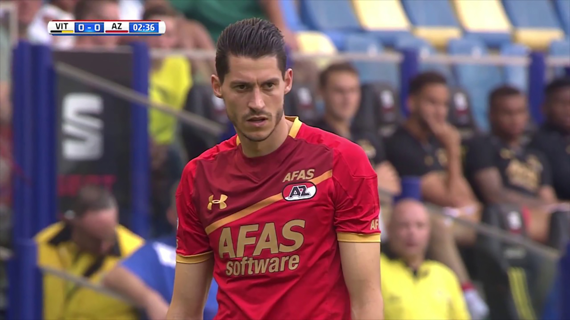 Voetballer in rood AZ-merkshirt op het veld, tegen een achtergrond van zittende toeschouwers en de score 0-0 tussen Vitesse (VIT) en AZ na 2 minuten en 36 seconden op het scherm.