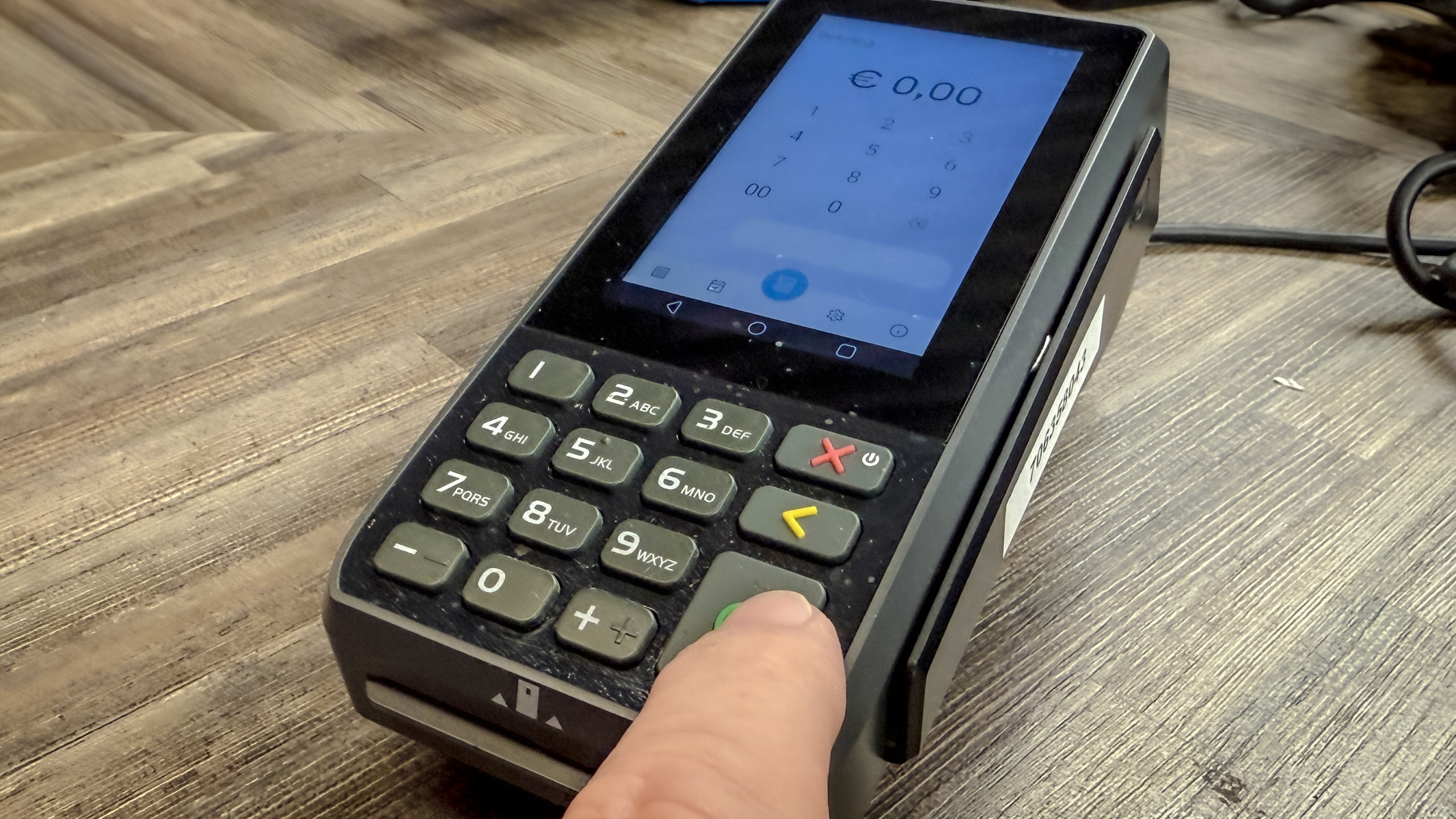Een betaalterminal met een scherm waarop €0,00 wordt weergegeven; er is een hand die een groene knop indrukt.