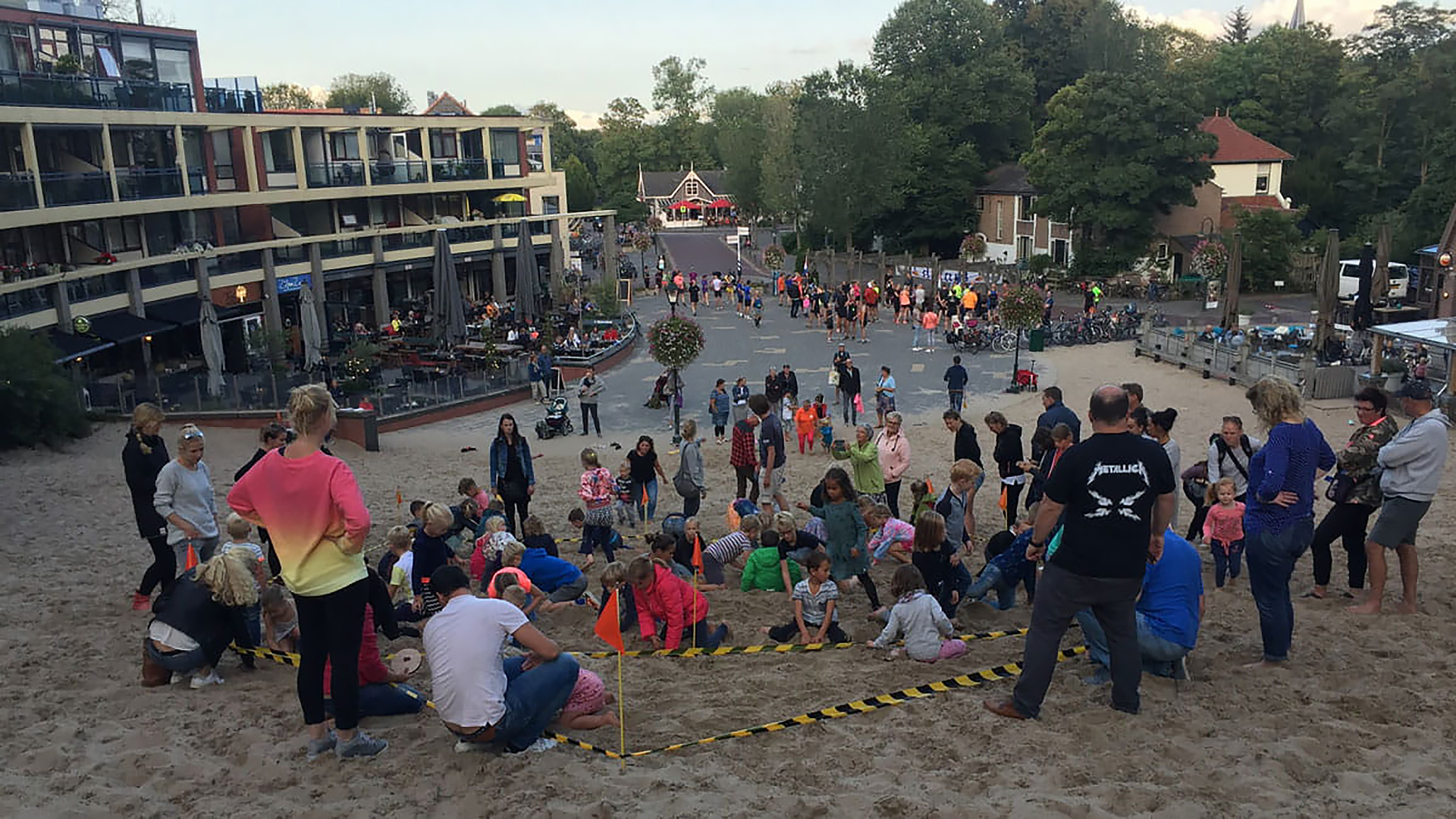 Groep mensen, waaronder veel kinderen, verzameld op een zandhelling voor een evenement, met een gebouw en terras op de achtergrond en een straat omringd door bomen verder weg.