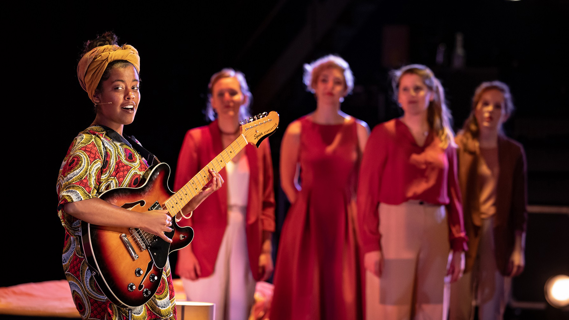 Vrouw met een elektrische gitaar, gekleed in kleurrijke kleding en een gele hoofdband, terwijl op de achtergrond vier andere vrouwen in roodachtige outfits staan.