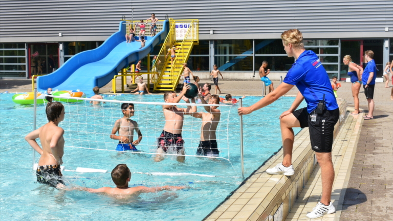 Kinderen spelen waterpolo in een buitenzwembad onder toezicht van een badmeester; op de achtergrond een glijbaan en andere zwembadactiviteiten.