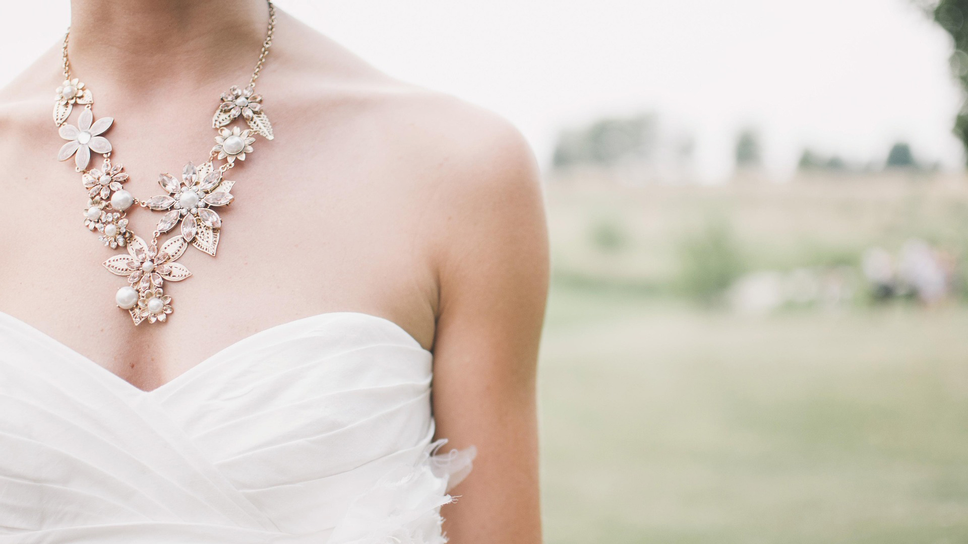 Close-up van een vrouw in een strapless witte jurk met een bloemrijke halsketting, op een buitenlocatie met wazige achtergrond.
