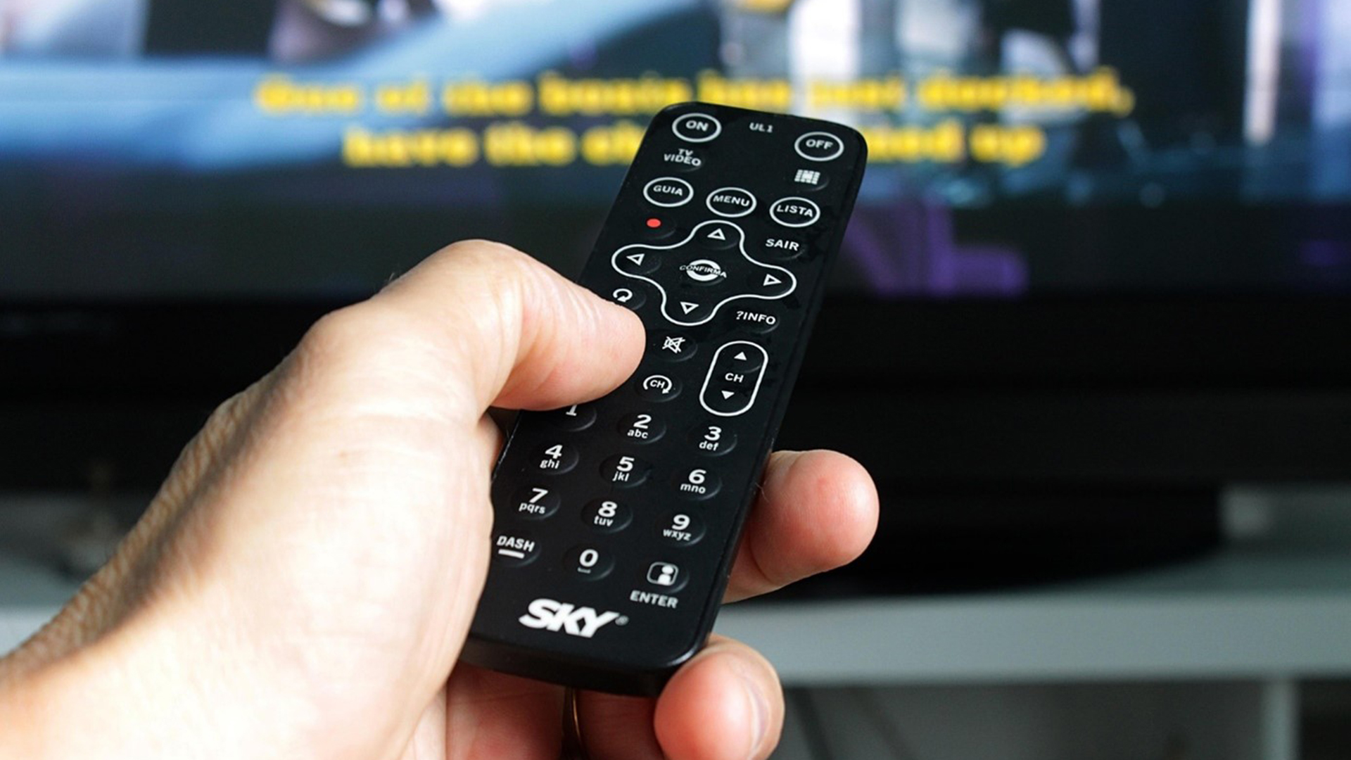 Hand die een televisie-afstandsbediening van Sky vasthoudt, gericht naar een televisiescherm met ondertiteling.
