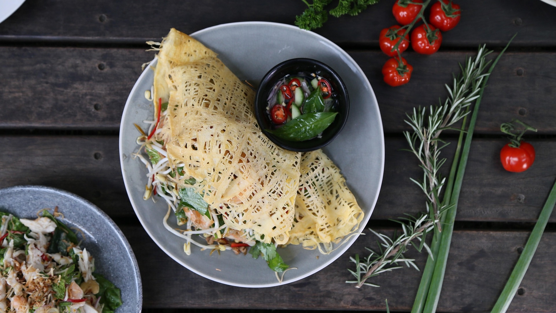 Een bord met een artistiek gevouwen gele crêpe gevuld met groenten, geserveerd met een klein zwart kommetje met dipsaus, omringd door verse kruiden en tomaatjes op een houten tafel.