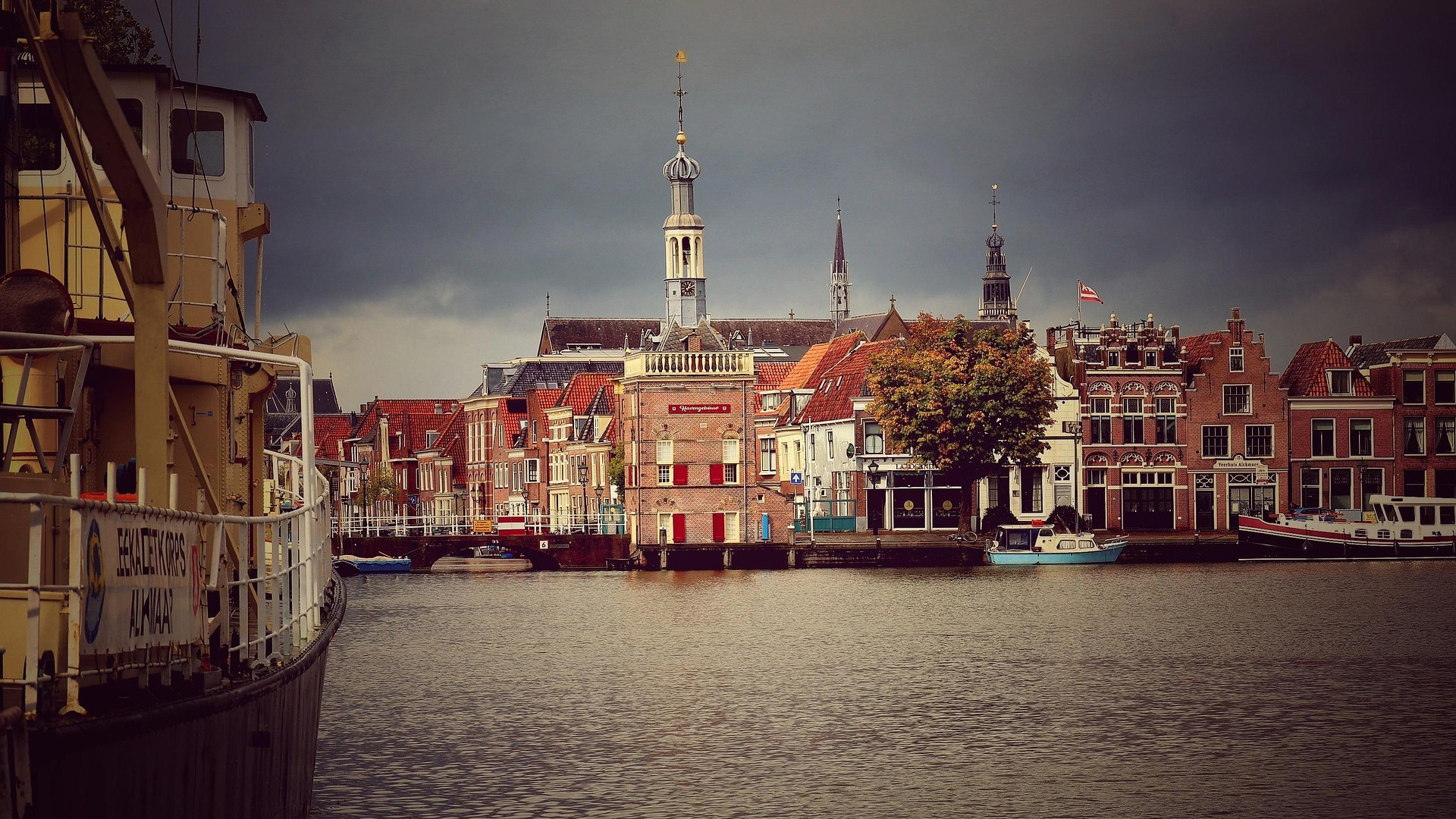Uitzicht op een haven met historische bakstenen gebouwen en schepen, met een torenspits en een bewolkte hemel op de achtergrond.