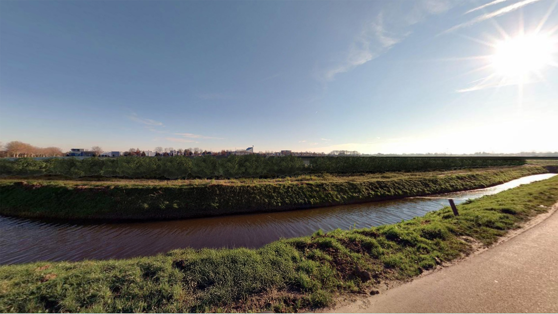 Landschap met een waterkanaal omringd door gras, en in de verte een rij huizen tegen een blauwe hemel met zonnige uitstraling.
