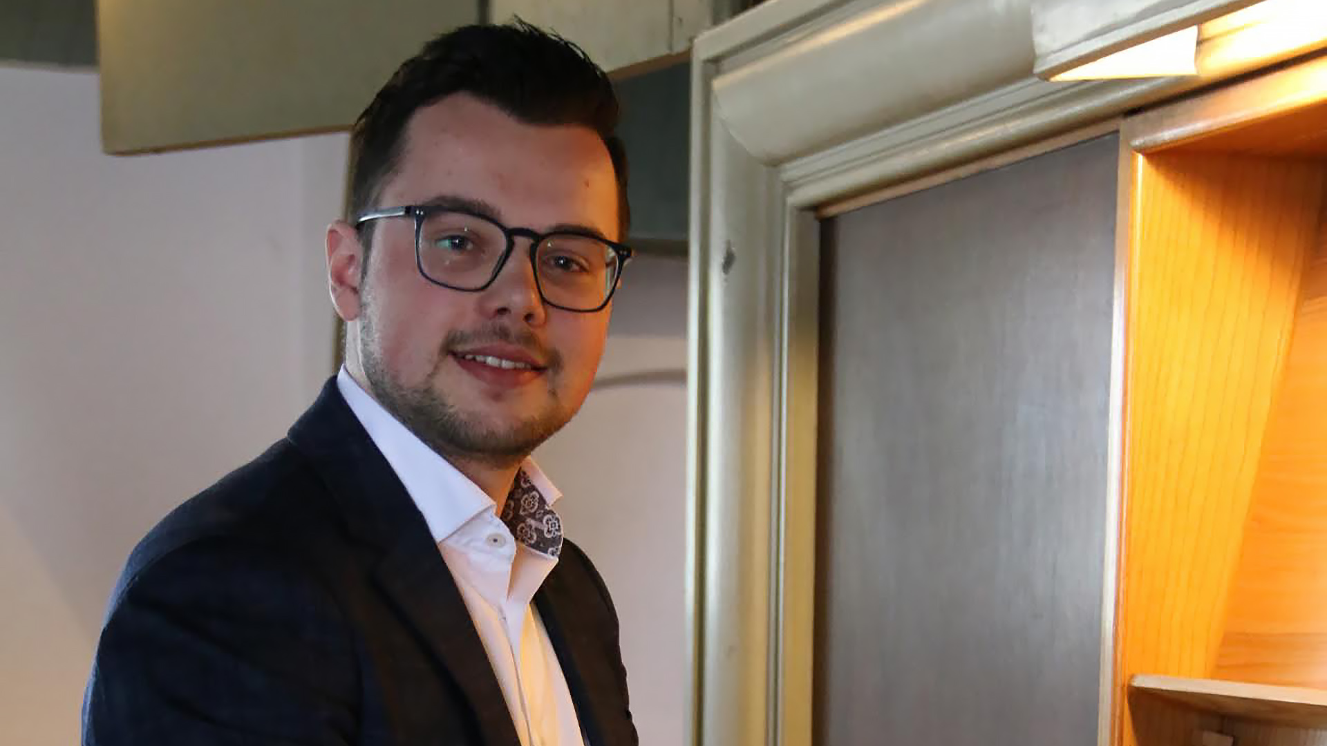 Een man met een bril draagt een donker pak en een witte blouse met een patroon in de kraag, terwijl hij in een kamer staat naast een open kast of plank.