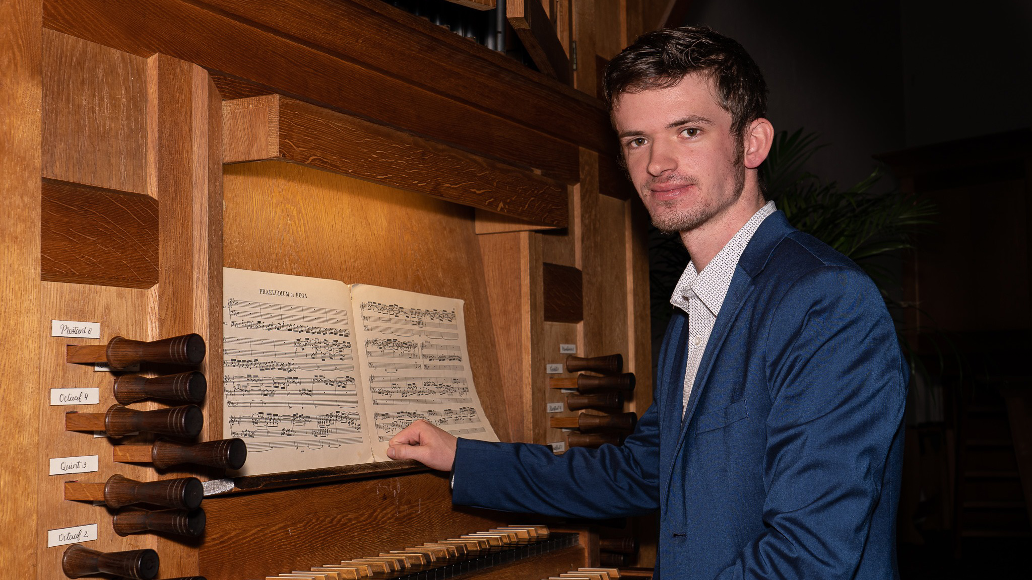 Een man in een blauwe colbert staat bij een kerkorgel, met muziekbladen geopend op de lessenaar en bedieningselementen zichtbaar aan de zijkant.