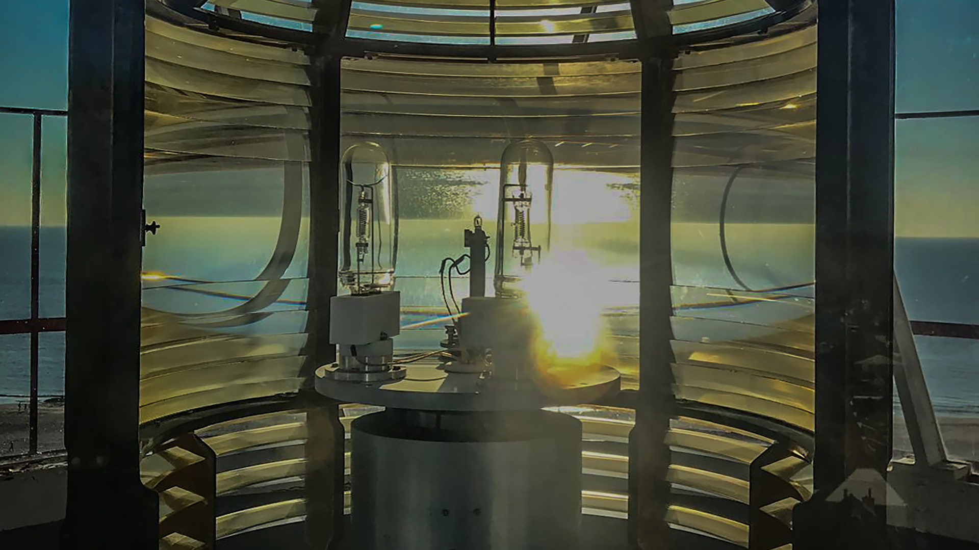 Binnenkant van een vuurtorenlens met zonlicht dat door de glazen panelen valt, met uitzicht op de oceaan.