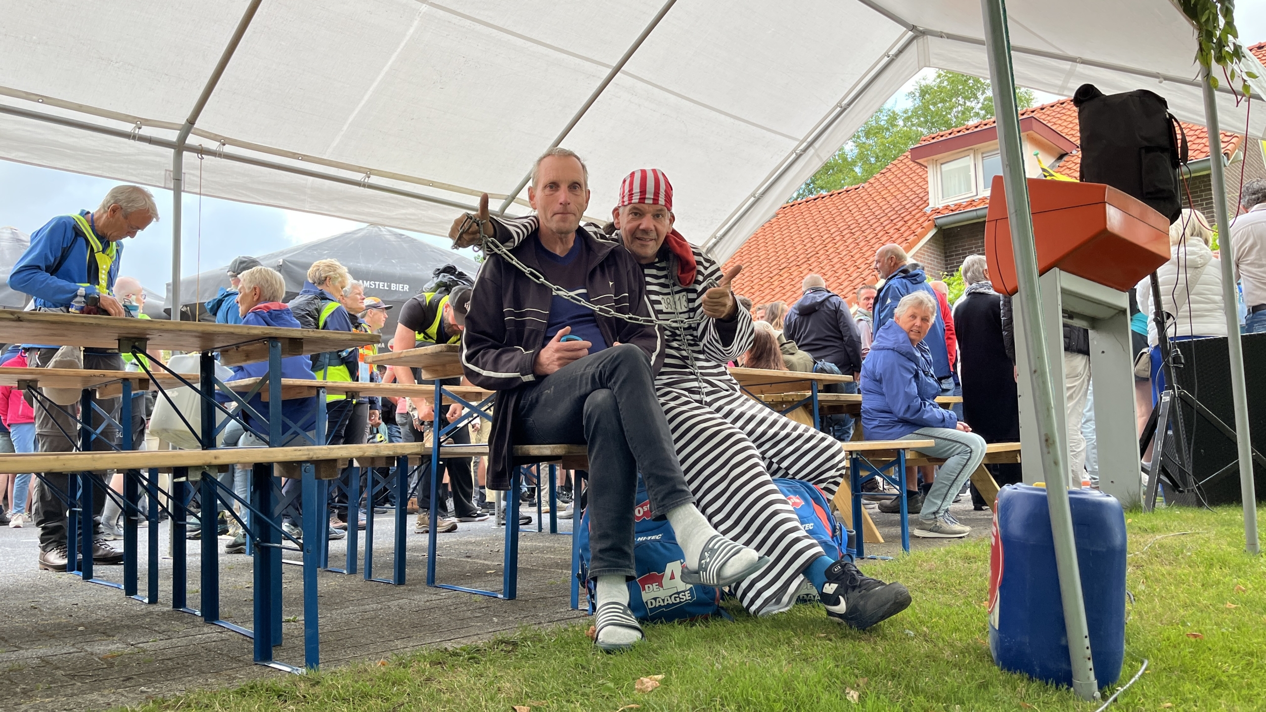 Twee mannen zitten op een bankje onder een tent, waarvan één opvallende zwart-wit gestreepte gevangeniskleding draagt en de andere is verkleed met een stok en kettingen om hen heen. Op de achtergrond staan mensen rond tafels bij een evenement.