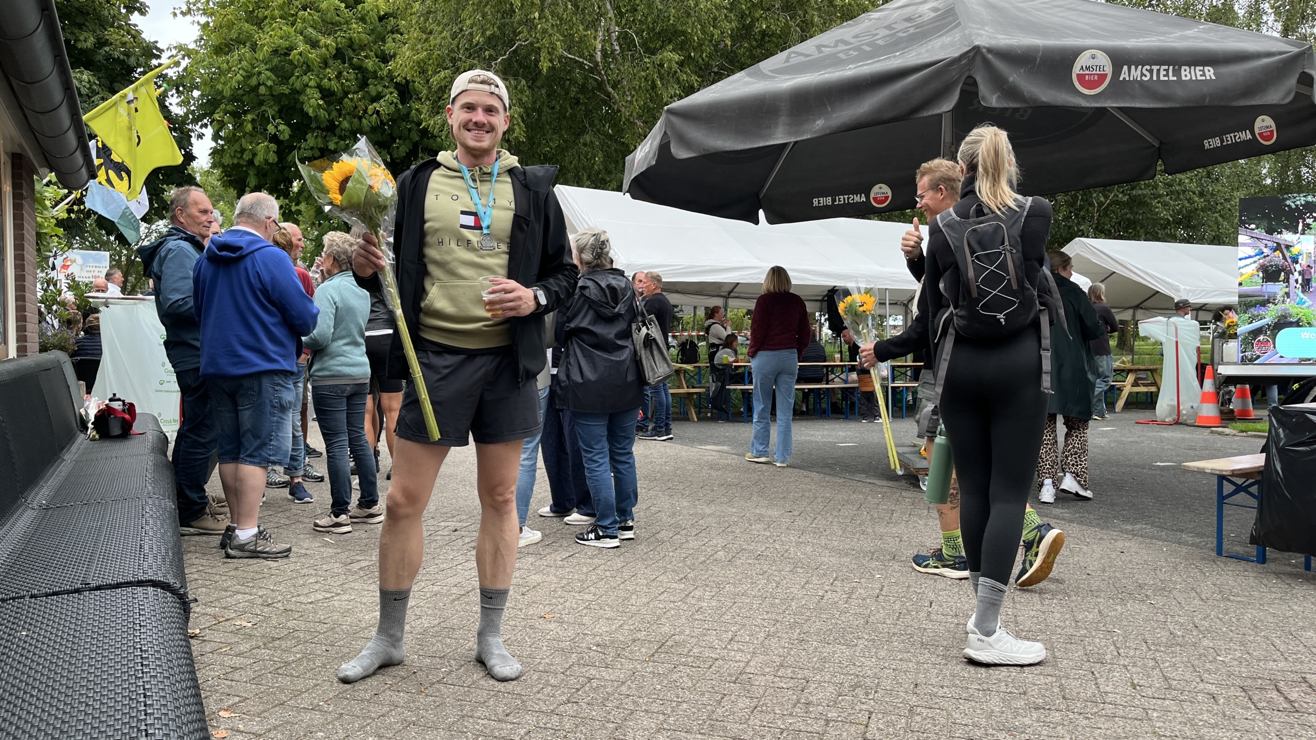 Een lachende man staat in een informele buitenbijeenkomst, draagt sportieve kleding en houdt een bos zonnebloemen vast. Op de achtergrond zijn andere mensen te zien onder grote Amstel Bier-parasols en tenten.