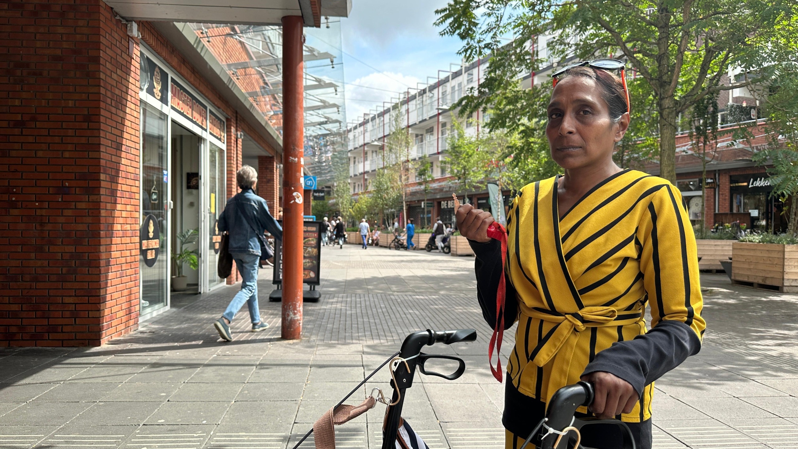 Een vrouw in een geel gestreept pak leunt op een rollator op een trottoir in een stadscentrum met bakstenen gebouwen en bomen op de achtergrond.