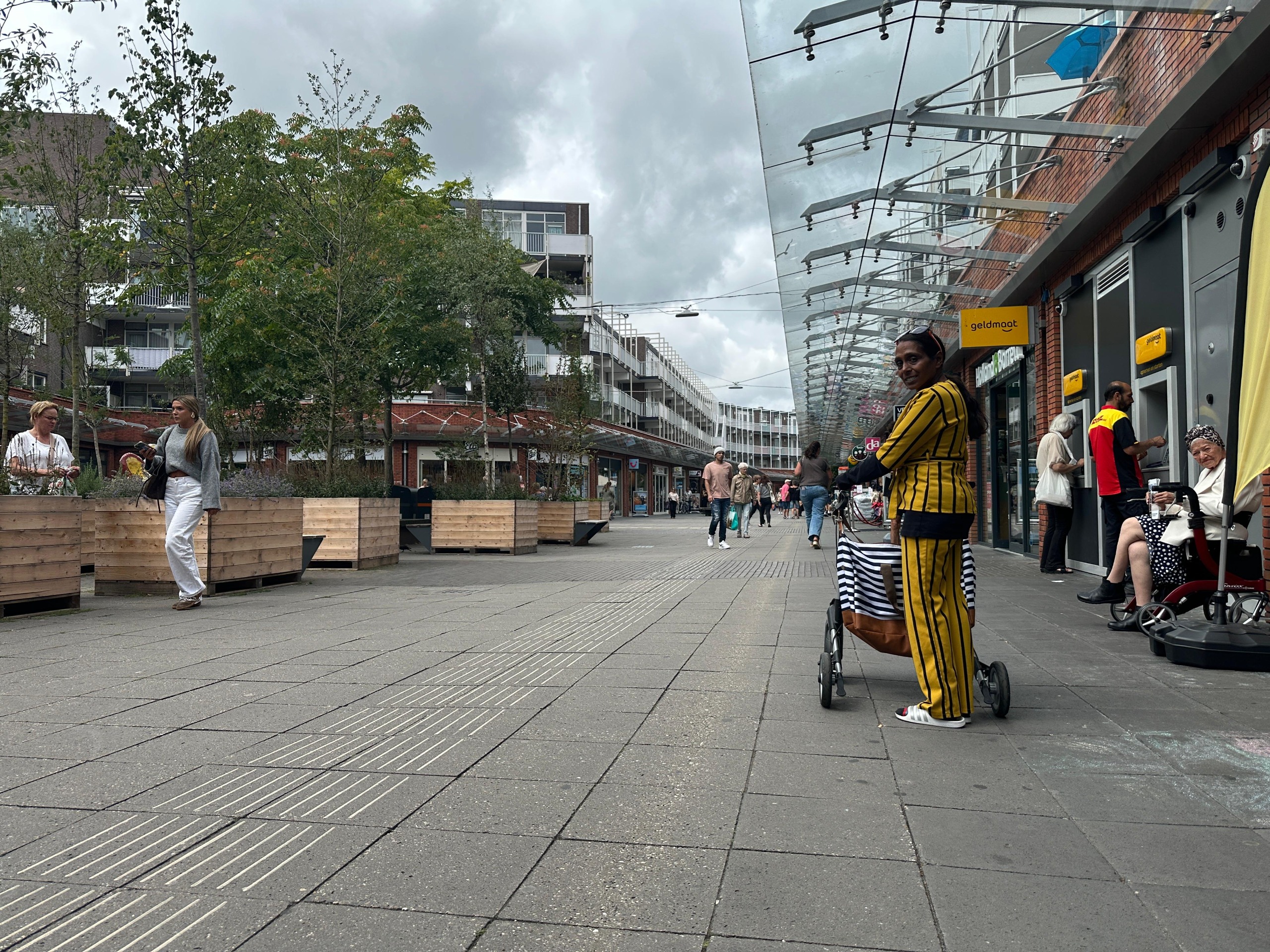 Straatscène in een winkelgebied met een vrouw in een geel gestreept pak die een kinderwagen duwt, omringd door andere voetgangers en winkels aan de zijkant.