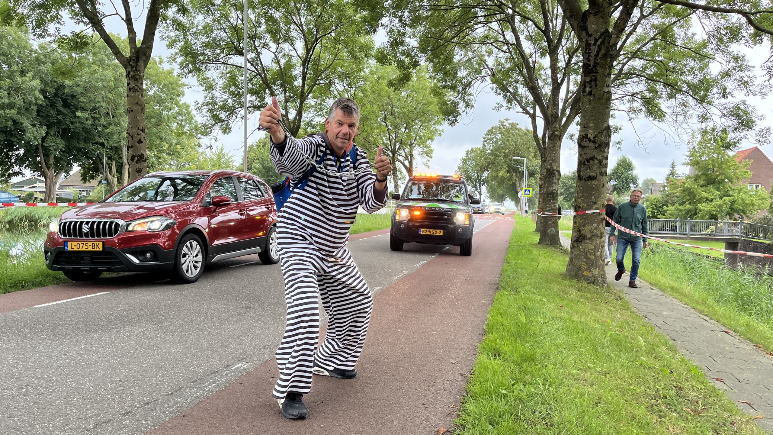 Man in een zwart-wit gestreept kostuum poseert op een weg met auto's en mensen op de achtergrond.