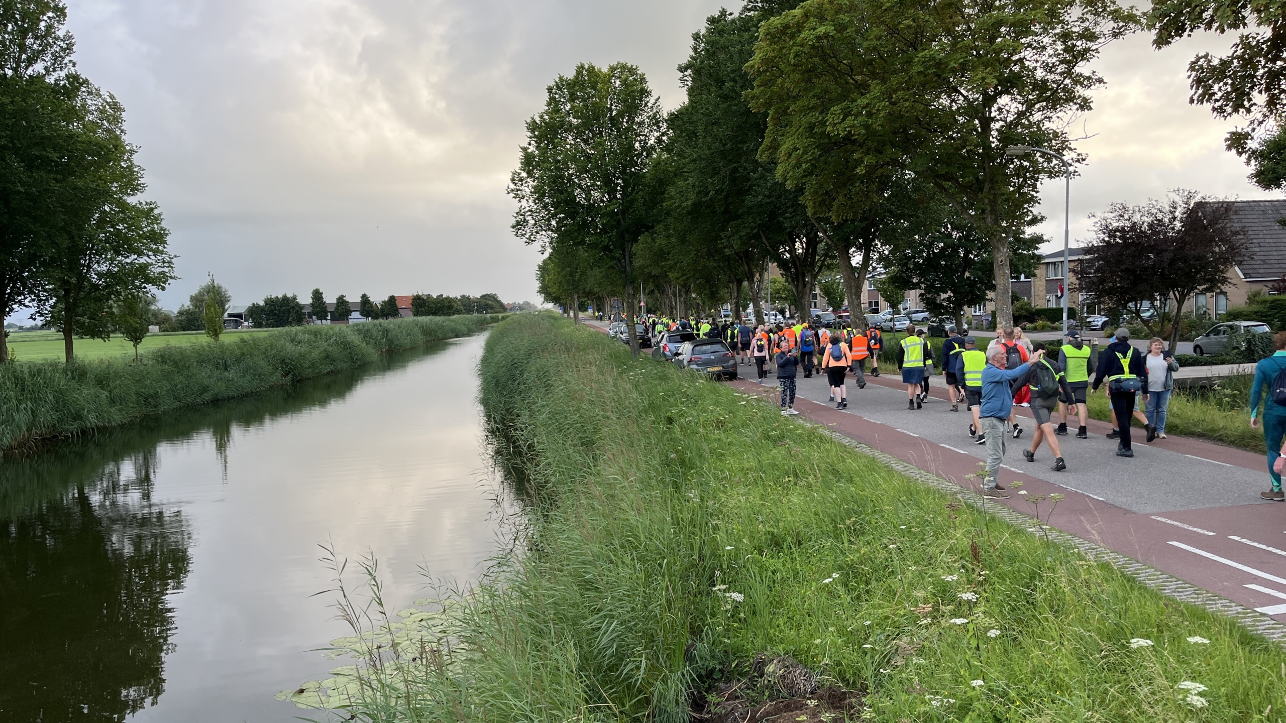 Mensen wandelen langs een kanaal bij een weg met rij bomen, gekleed in sportkleding en veiligheidsvesten.