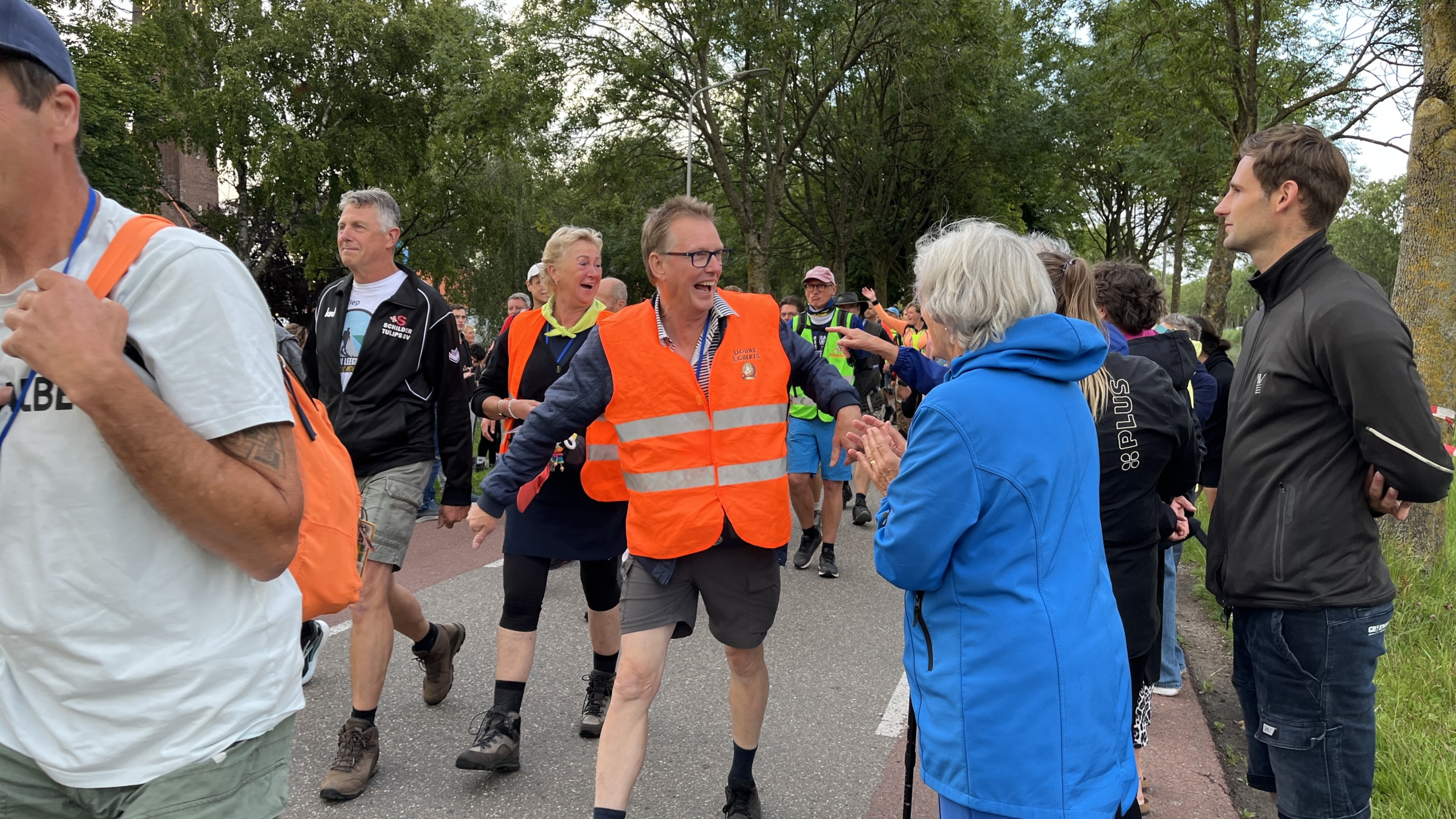 Mensen lopen in een groep over een weg, met één persoon in een fel oranje veiligheidsvest die lachend contact maakt met toeschouwers langs de kant. Er zijn meerdere mensen in sportieve kleding en verschillende achter hen marcherende figuren, tegen een achtergrond van bomen en een heldere lucht.