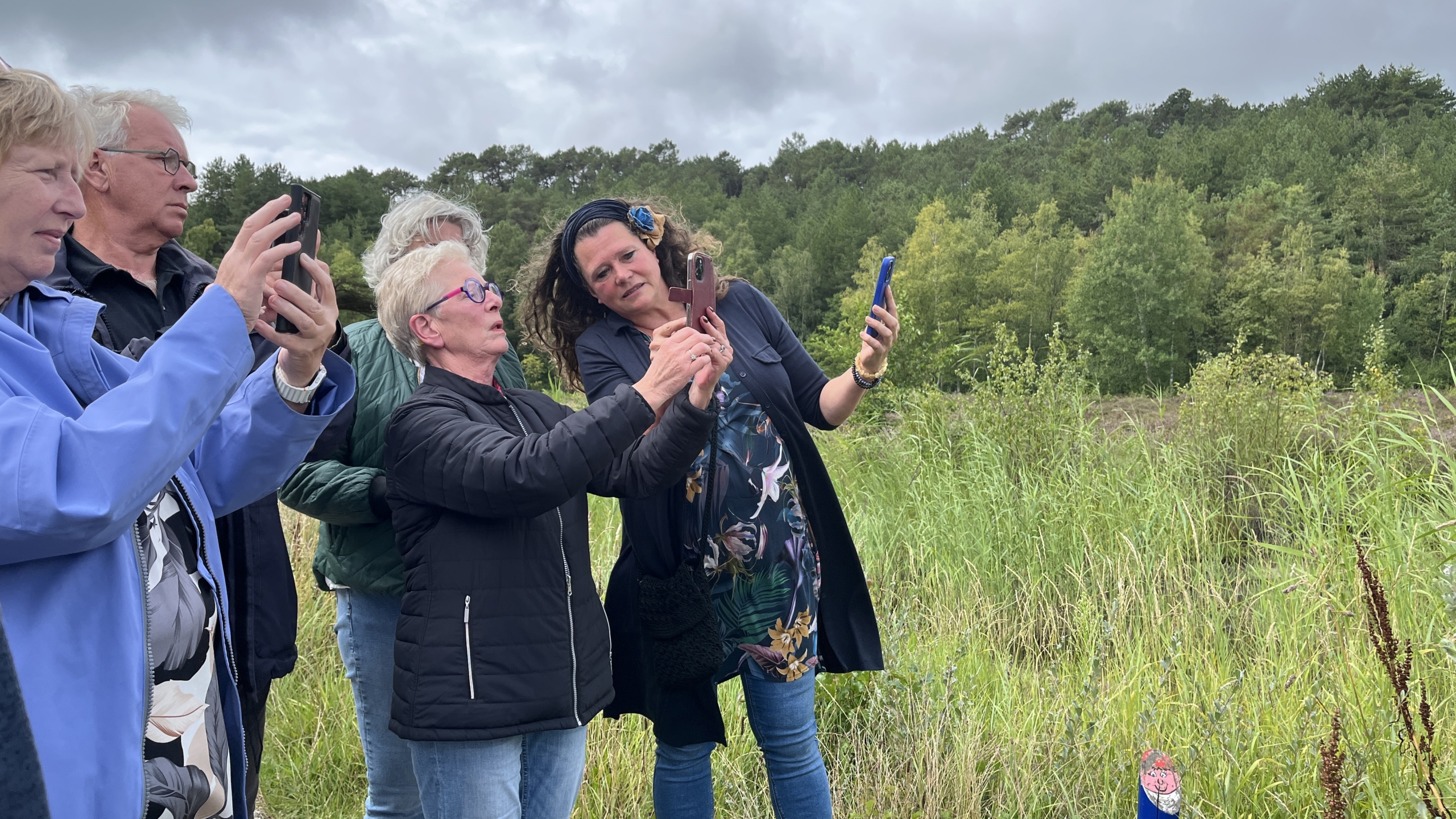 Een groep mensen staat in een veld met gras en bomen op de achtergrond, terwijl ze met hun telefoons foto's maken of iets bekijken.