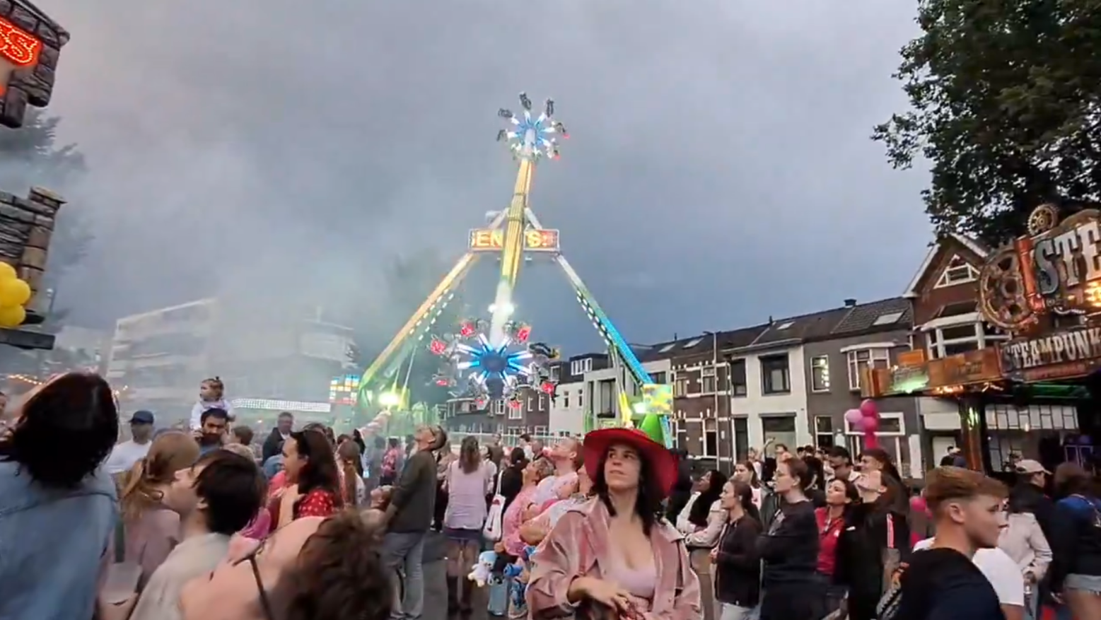 Mensen verzamelen zich op een drukke kermis, met een grote kermisattractie op de achtergrond die kleurrijke lichten en bewegingen heeft. De lucht is bewolkt, en er zijn diverse stands en kraampjes zichtbaar.