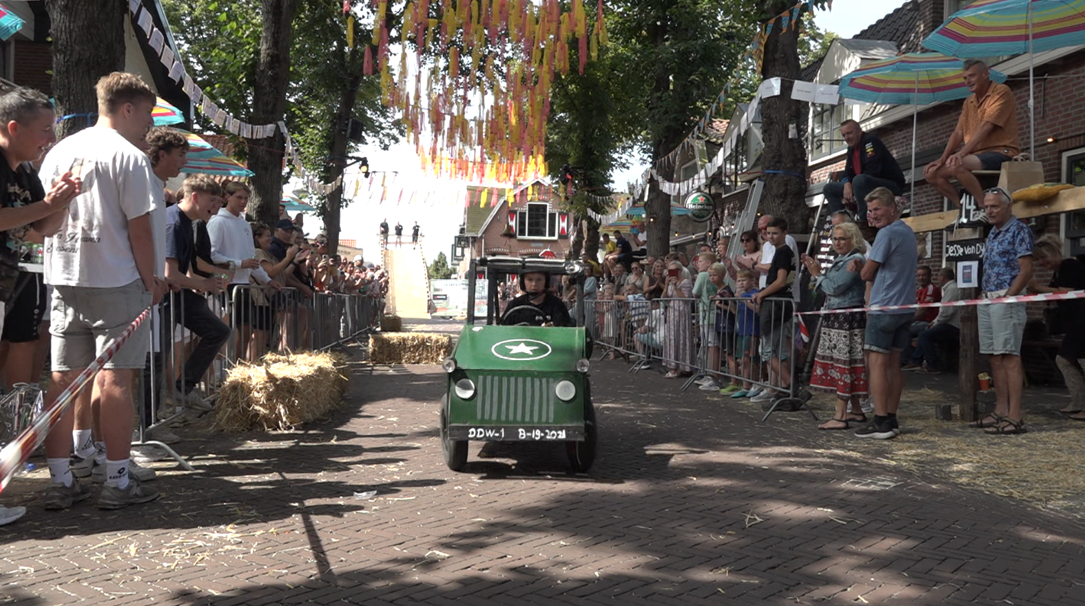 Een kind rijdt in een groen speelgoedvoertuig dat lijkt op een jeep door een straat met toeschouwers aan beide kanten, omringd door bomen en versieringen.