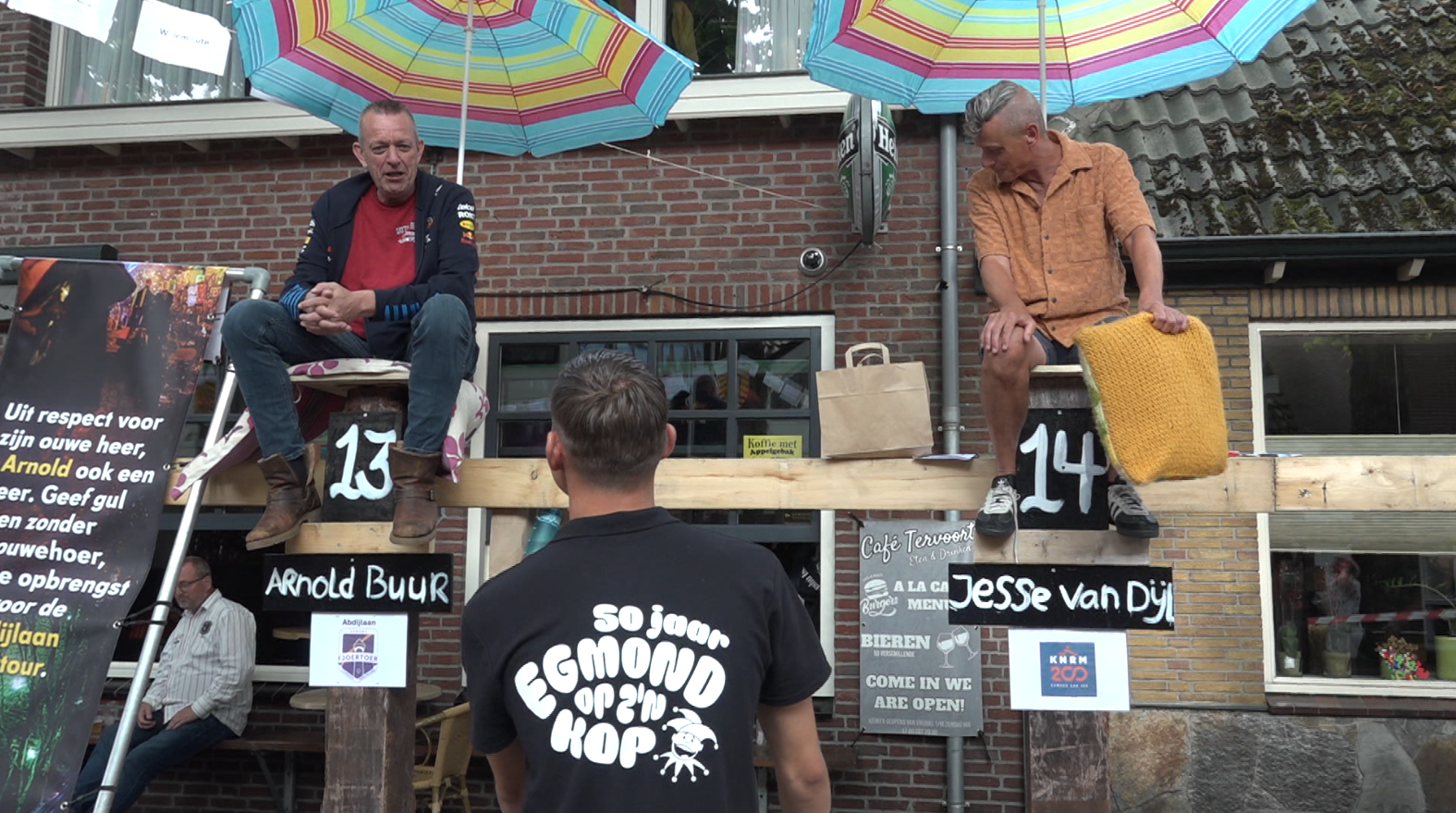 Twee mannen zitten op houten balken met nummers 13 en 14, onder kleurrijke parasols, tijdens een evenement buiten een café, terwijl een andere man hen aankijkt.