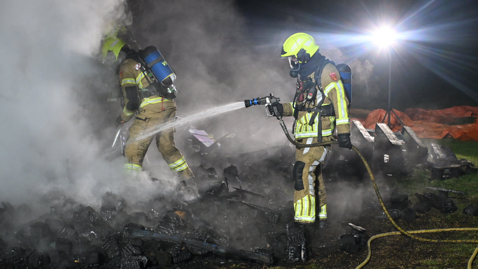 Brandweerlieden bestrijden een brand met waterstralen in de nacht, te midden van dikke rook en verlichte omgeving.