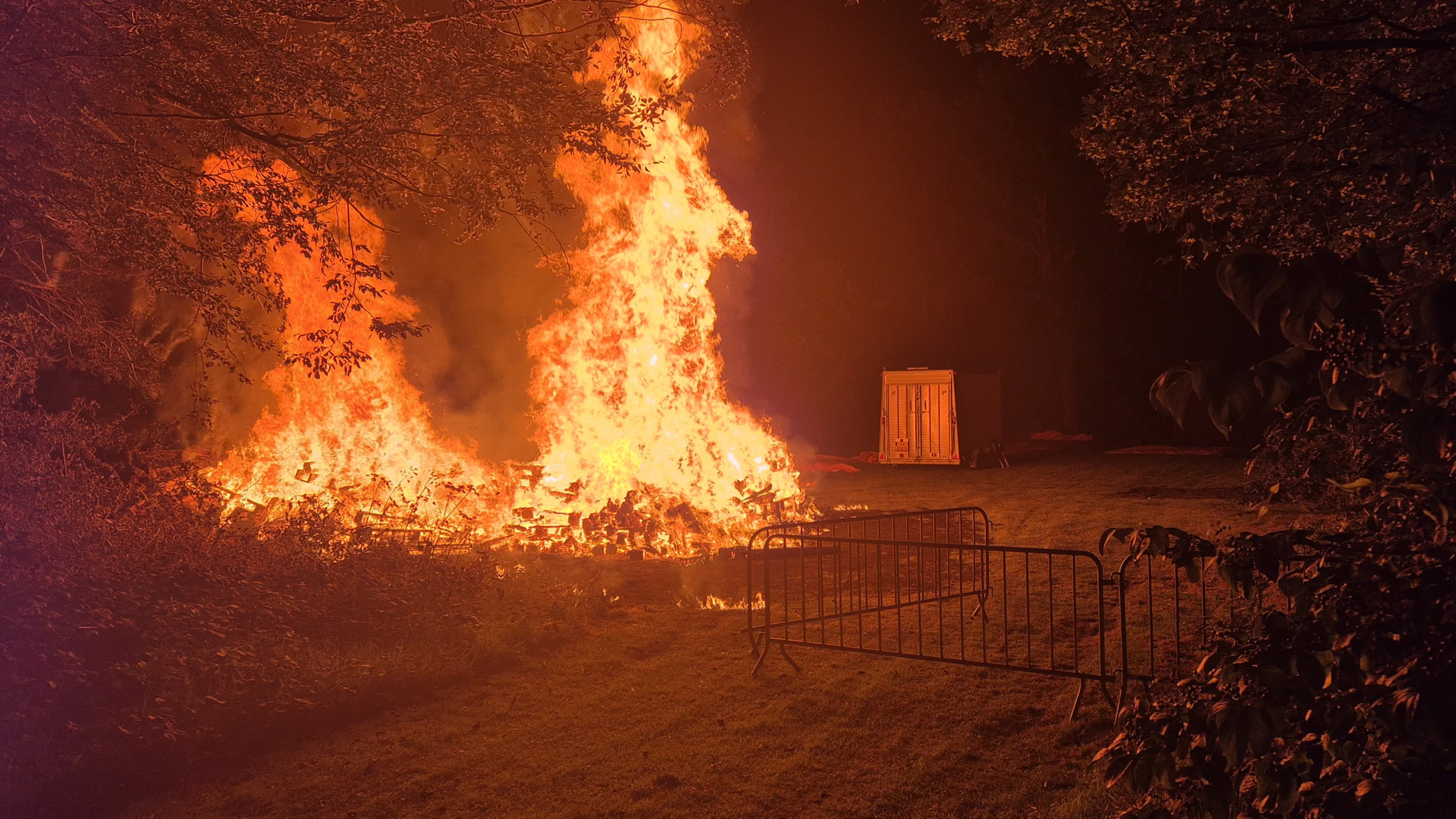 Brandende stapel hout met hoge vlammen, omringd door bomen en een metalen hek, in een nachtelijke omgeving.