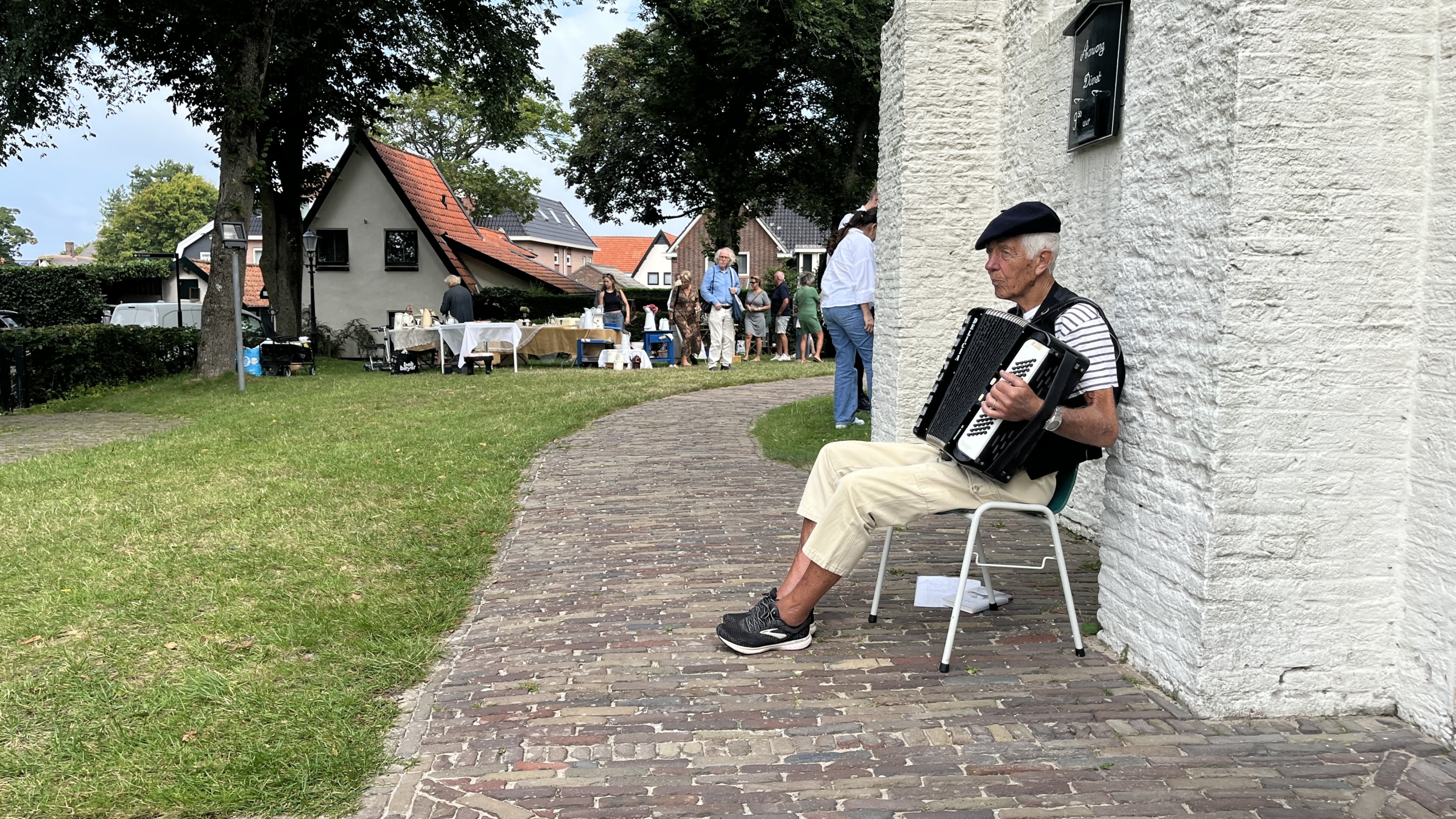 Een oudere man speelt accordeon zittend op een stoel naast een wit stenen gebouw, terwijl in de achtergrond mensen door een straatje wandelen langs kraampjes met goederen.