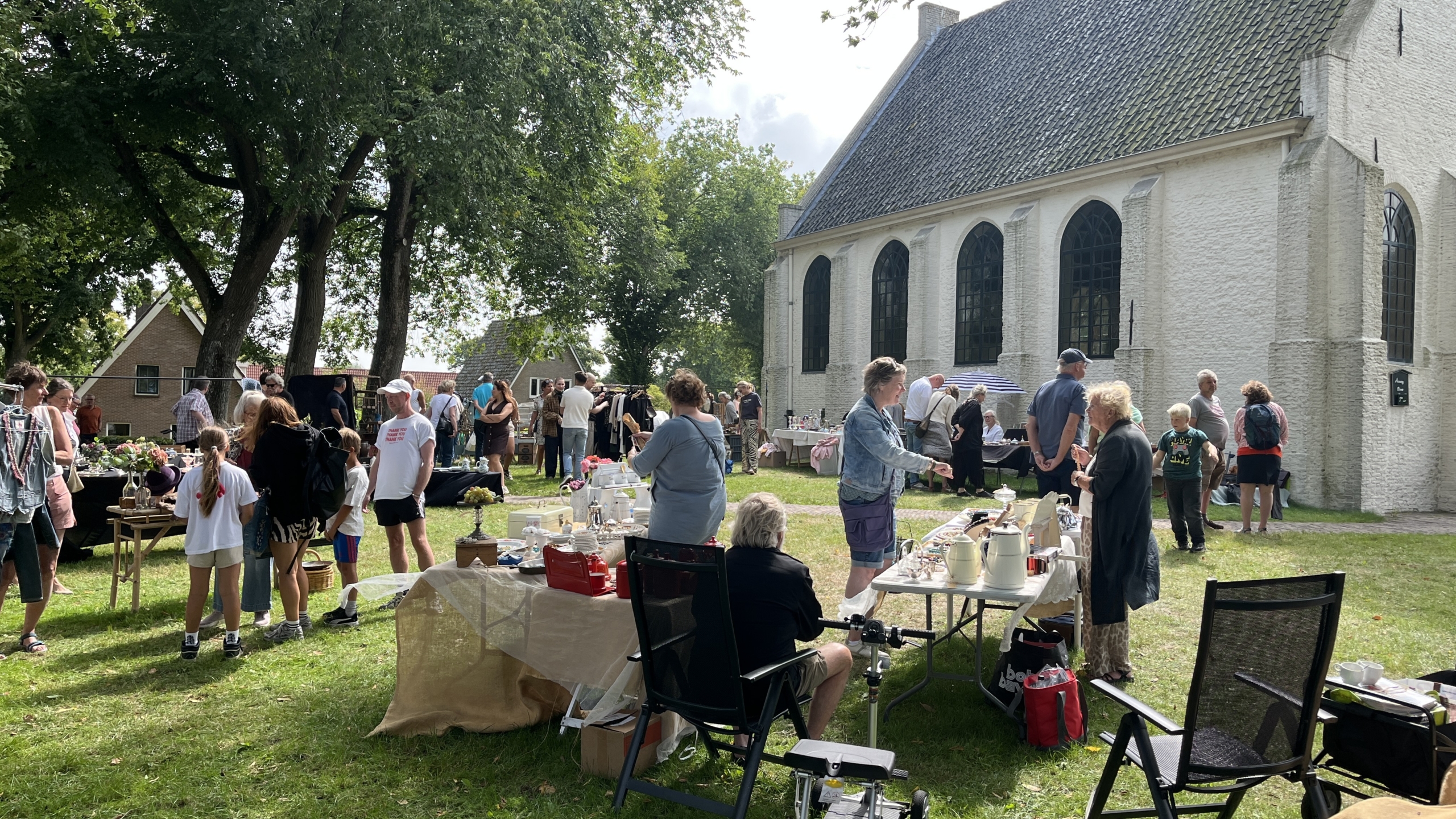 Een buitenmarkt met mensen die rondlopen en staan bij tafels met tweedehands goederen, naast een witte kerk met hoge ramen.
