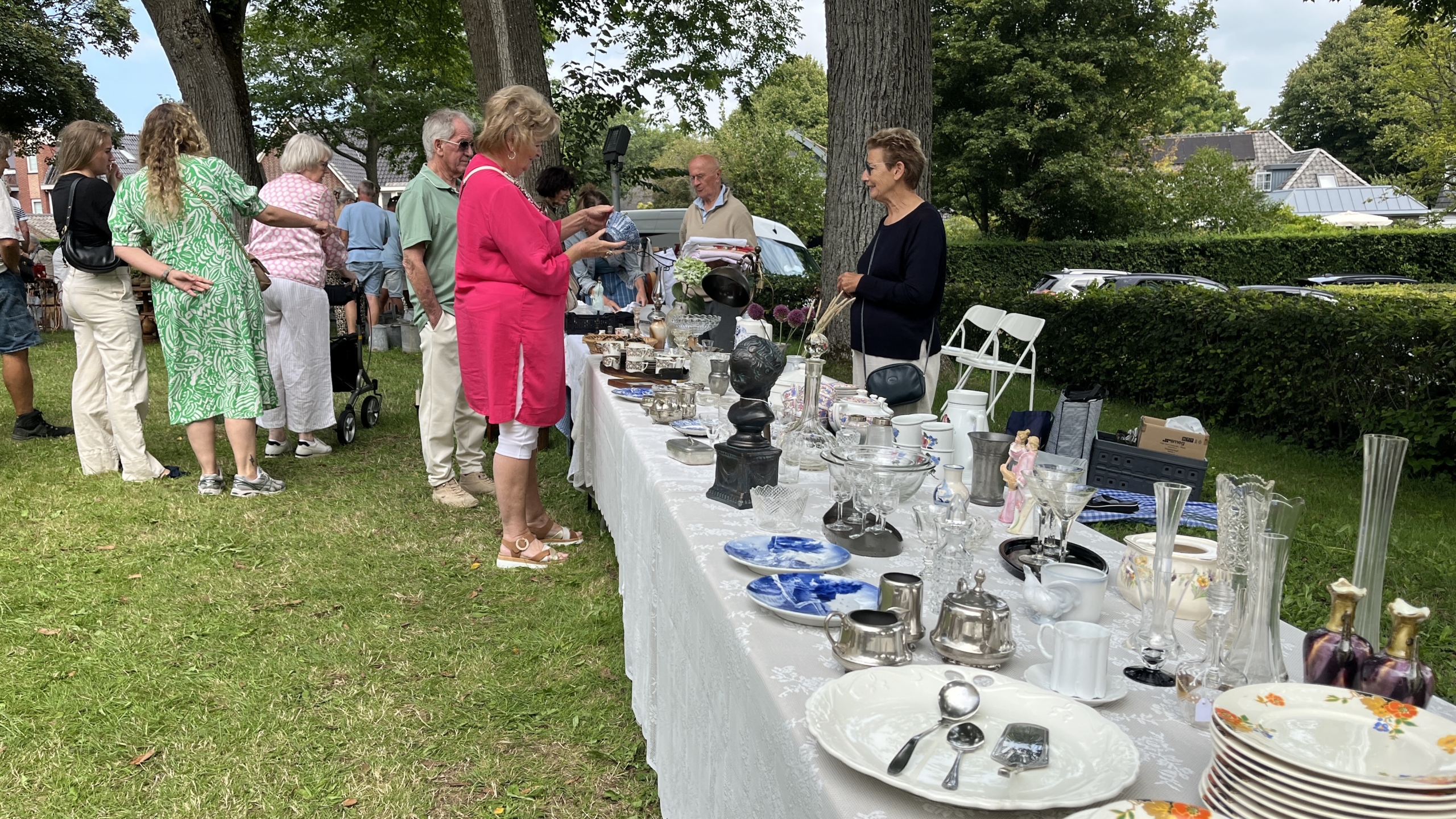 Mensen bekijken en kopen antiek en curiosa op een rommelmarkt met tafels vol serviesgoed, vazen en decoratieve objecten, omringd door bomen en grasvelden.