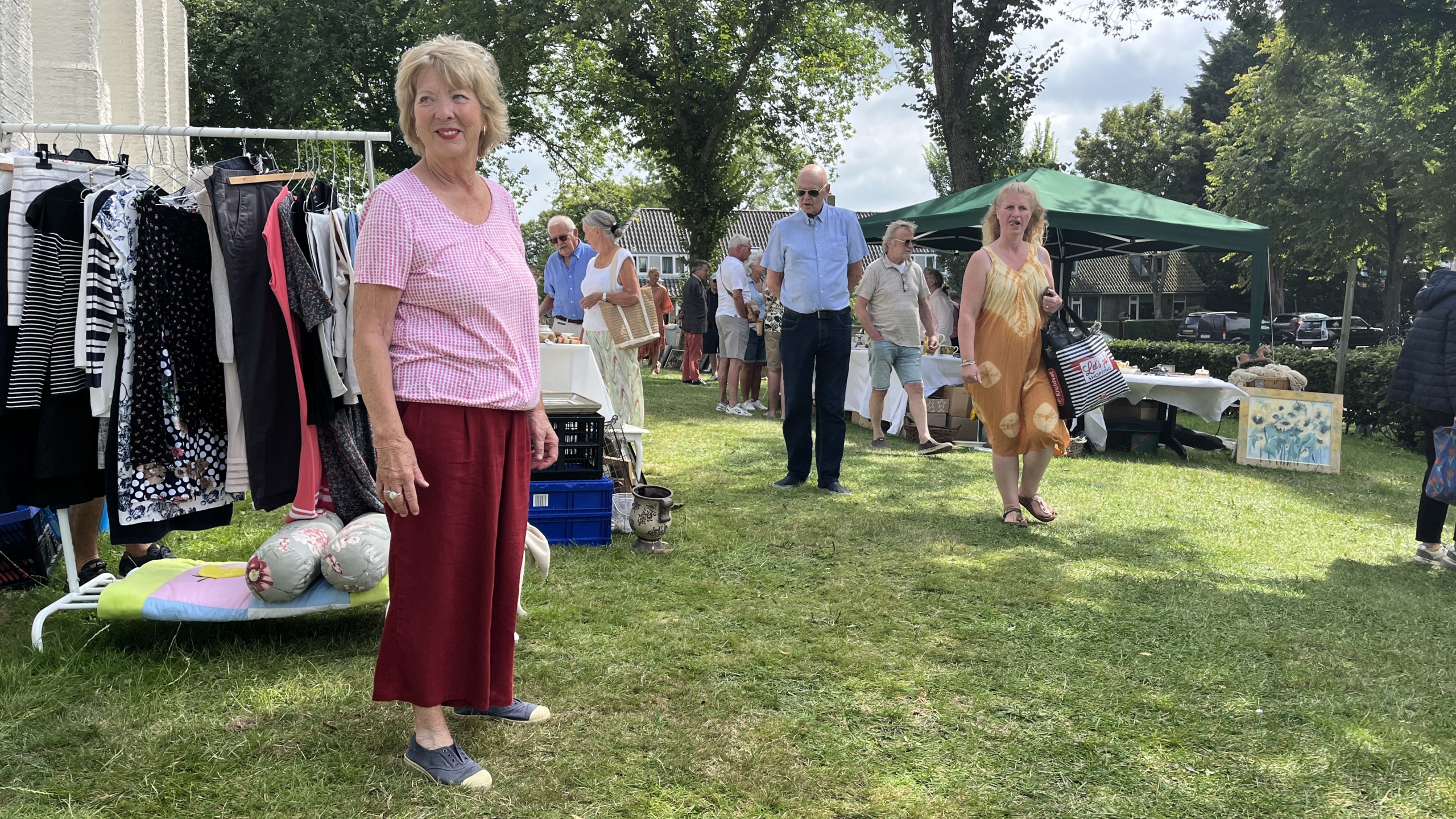 Een buitenmarkt op gras met een vrouw in een roze geruite blouse en rode broek die bij een kledingrek staat, omringd door mensen bij verschillende kraampjes.