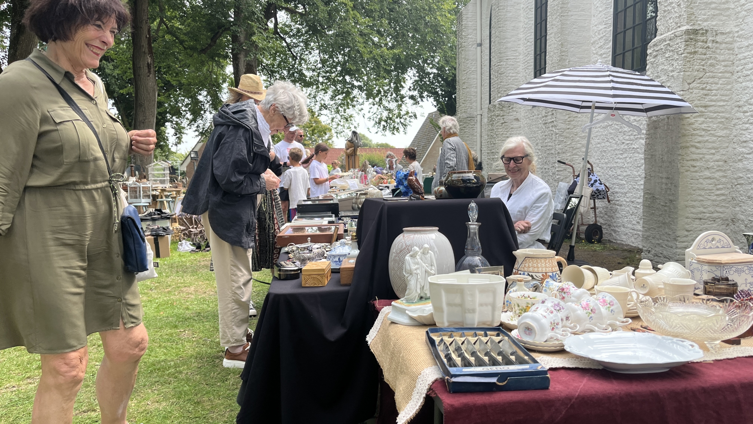 Mensen bekijken en verkopen antieke en vintage items op een buitentafelmarkt naast een stenen gebouw met ramen.