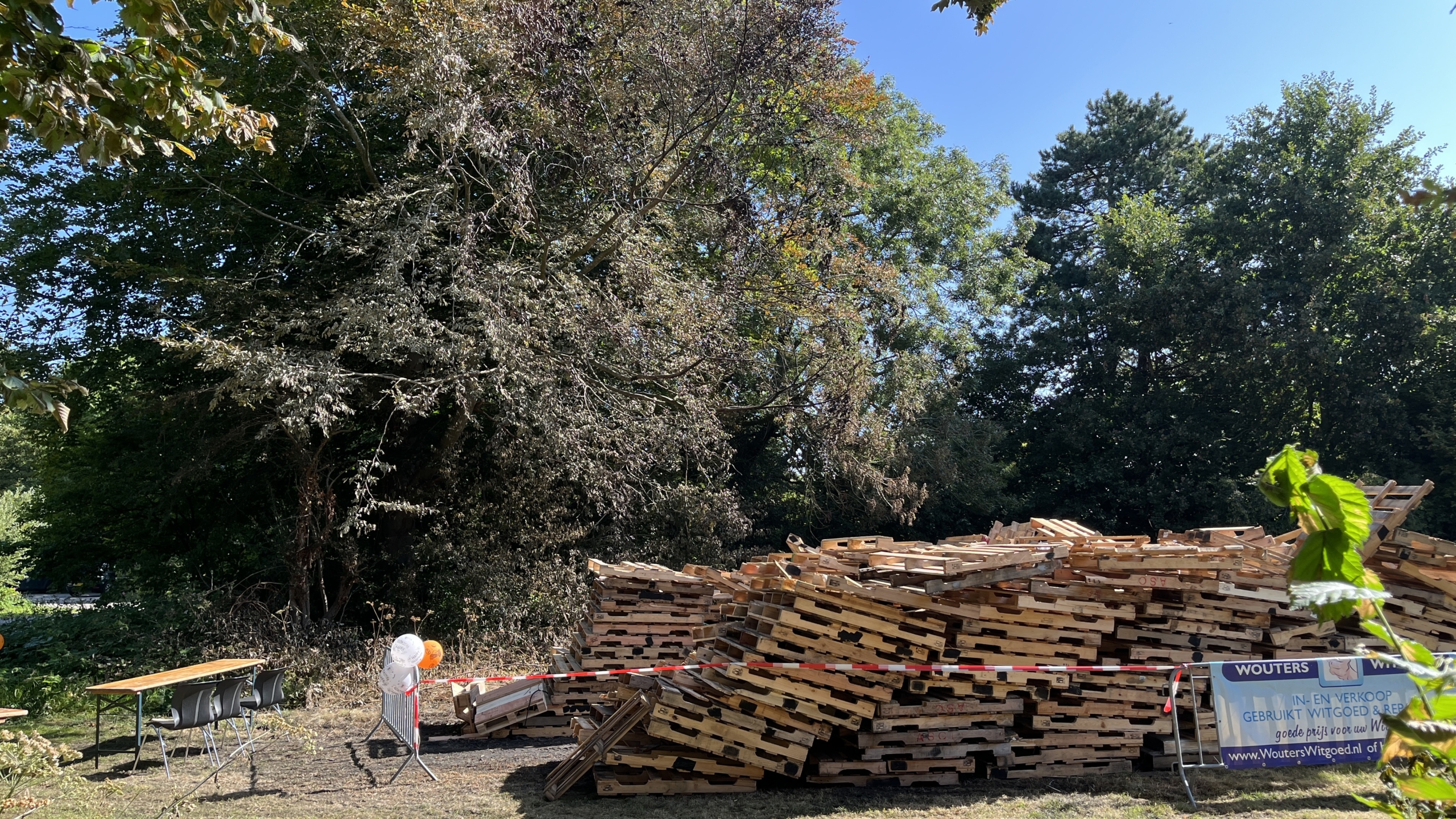 Een grote stapel houten pallets staat op een grasveld naast bomen, met een reclamebord en een tafel met stoelen in de buurt.