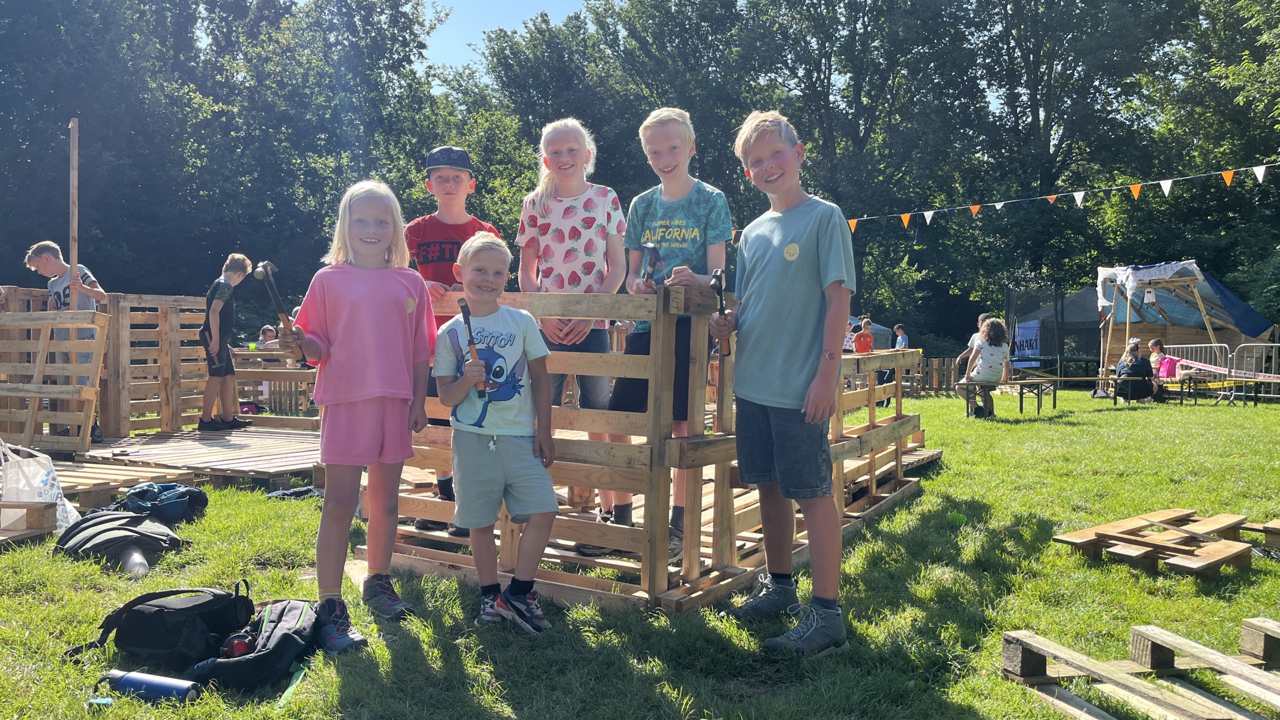 Kinderen poseren met hamers voor een houten bouwwerk op een zonnig grasveld, omringd door palletten en spelende kinderen in de achtergrond.