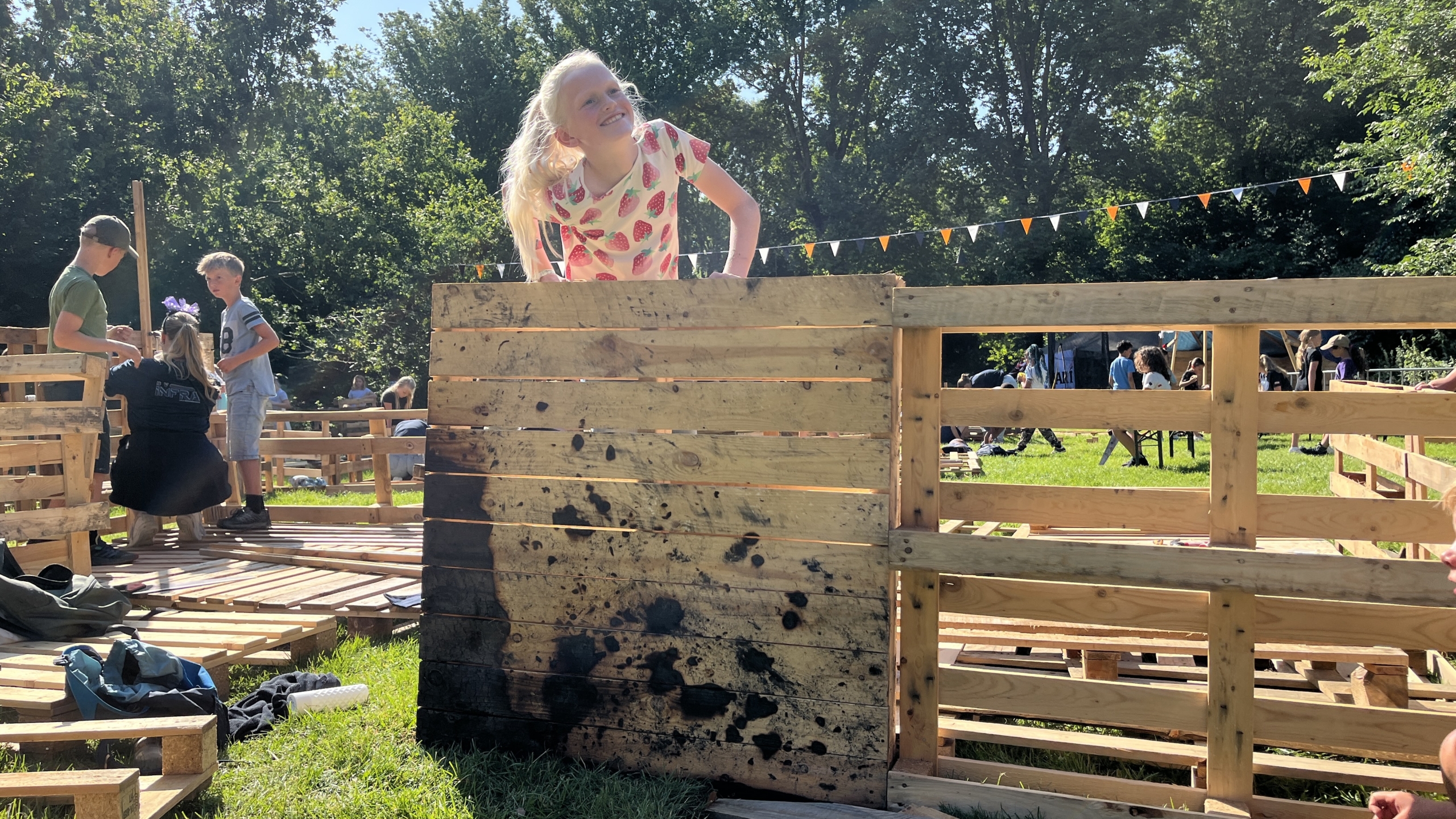 Kinderen bouwen met houten pallets op een zonnige dag in een park, omringd door groen en kleurrijke vlaggetjes.